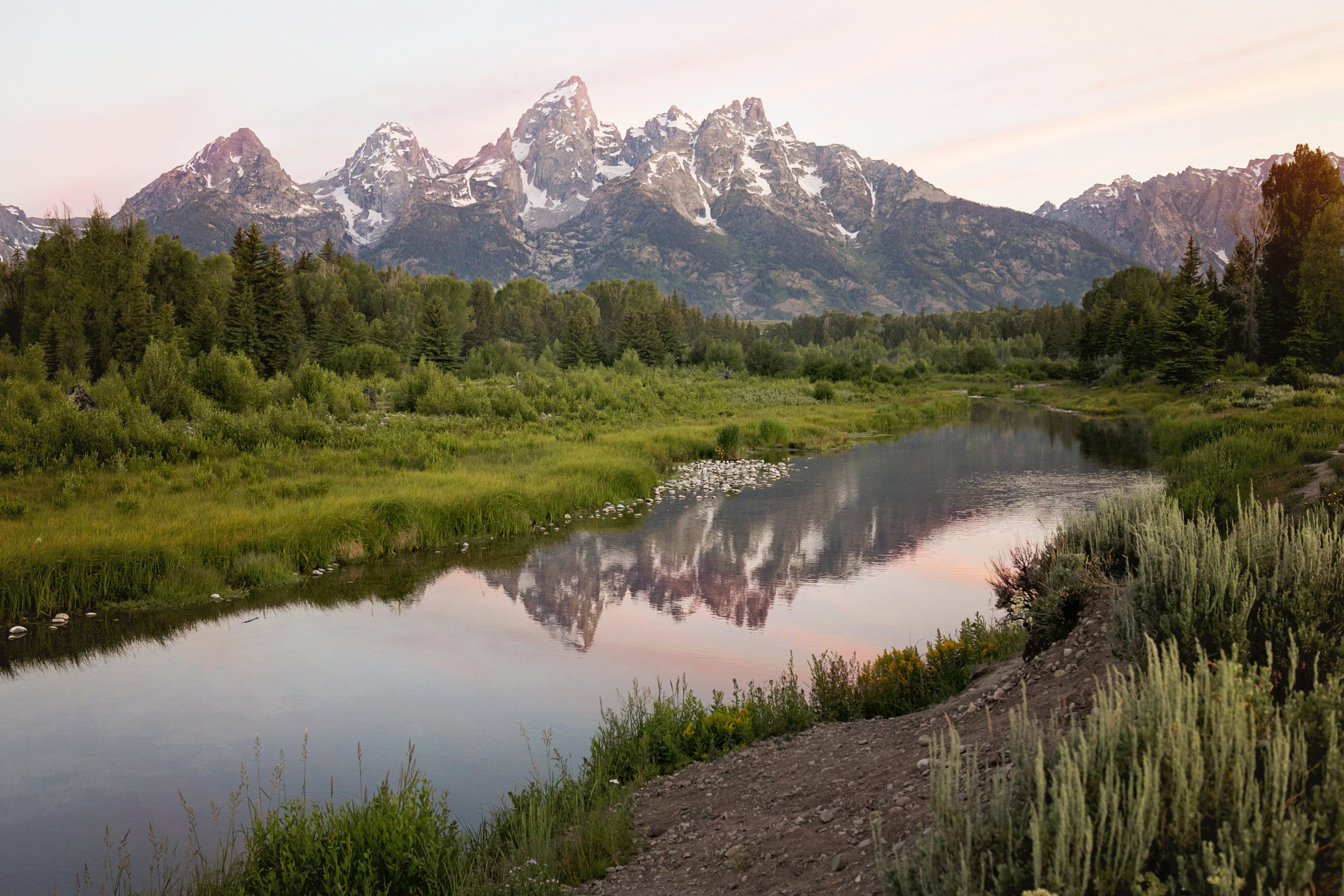 Mountain range with snow-capped peaks, lush green trees, a calm river reflecting the scenery, and a dirt path along the riverbank during sunset.