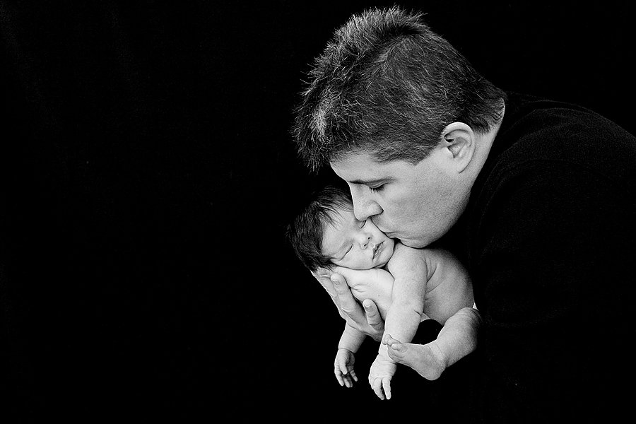 A man gently kisses a tiny, newborn baby on the forehead in a black-and-white photo.