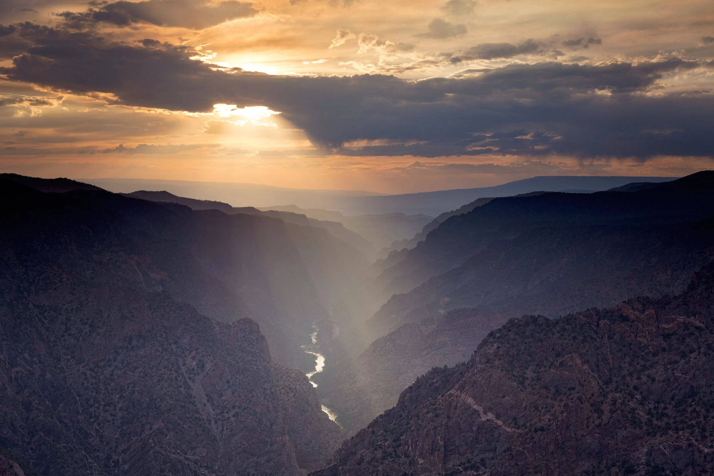 Sunset over a canyon with mountain ridges, cloudy sky, and sunlight breaking through clouds.