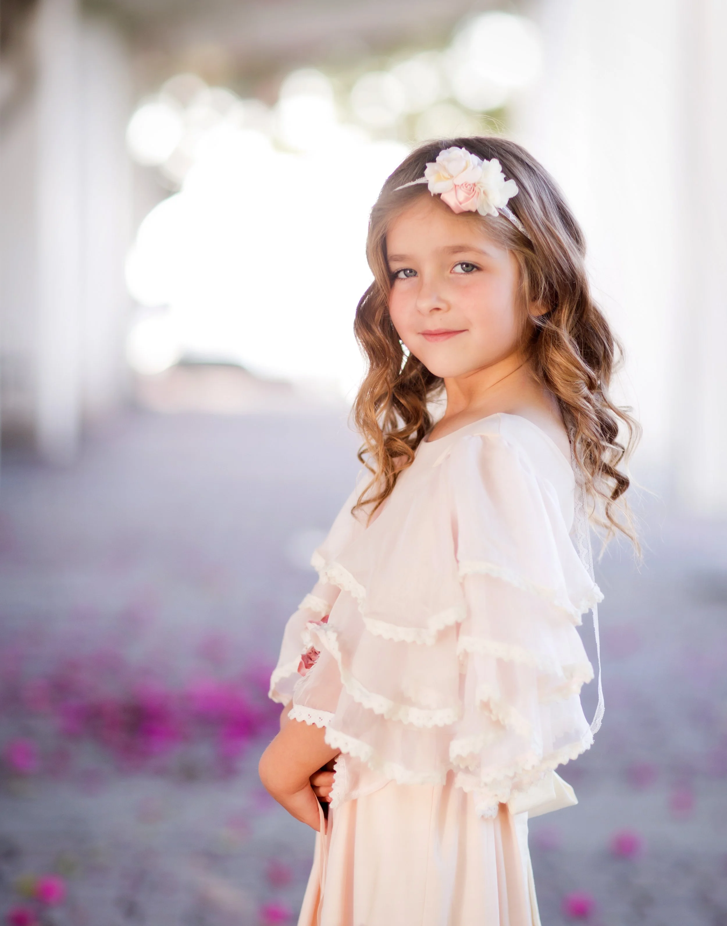 A young girl with wavy brown hair and blue eyes, wearing a light pink dress with lace ruffles and a floral headband with white and pink flowers, standing outdoors in a blurred background with bokeh of light.