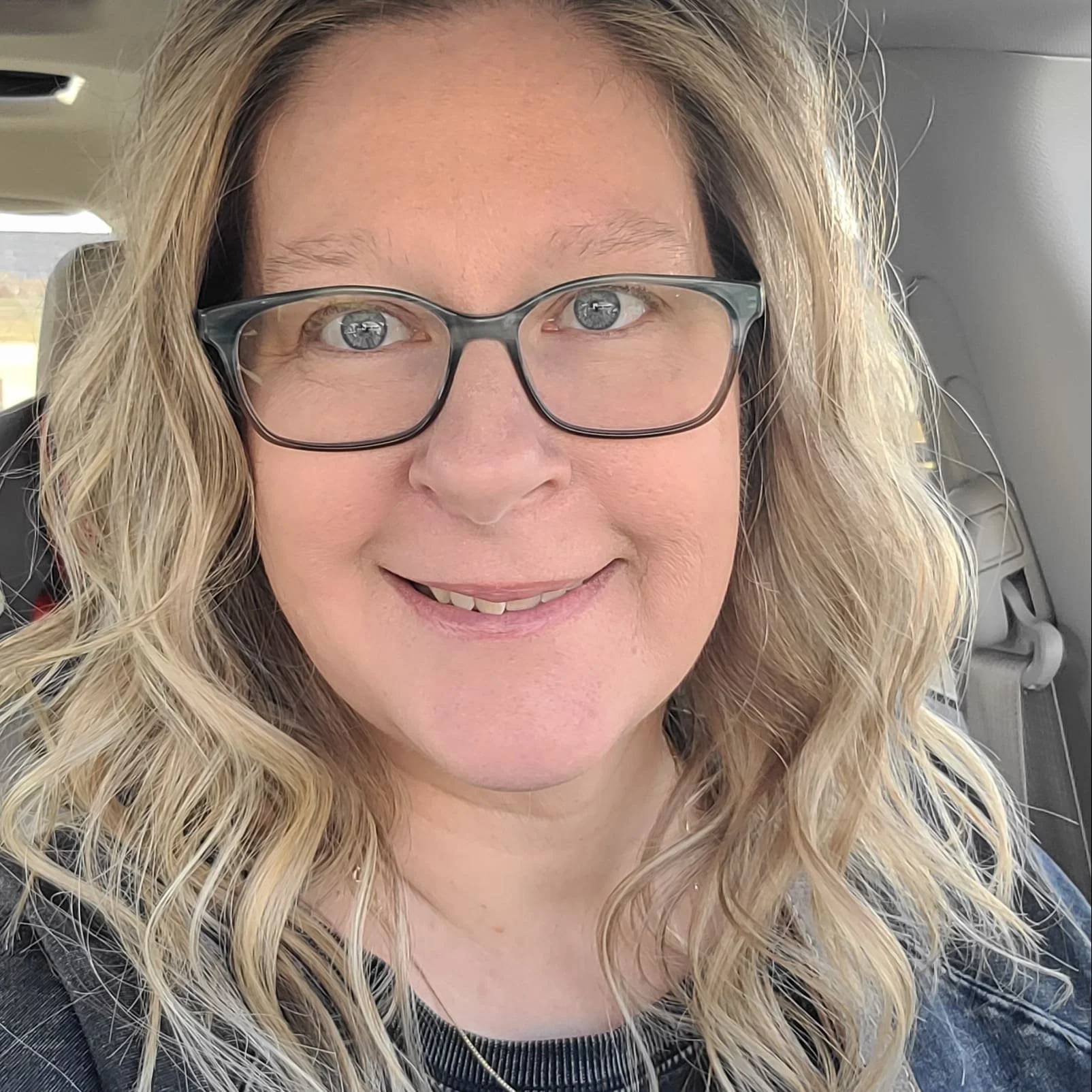 Close-up portrait of a woman with blonde, curly hair wearing glasses, smiling in a vehicle.