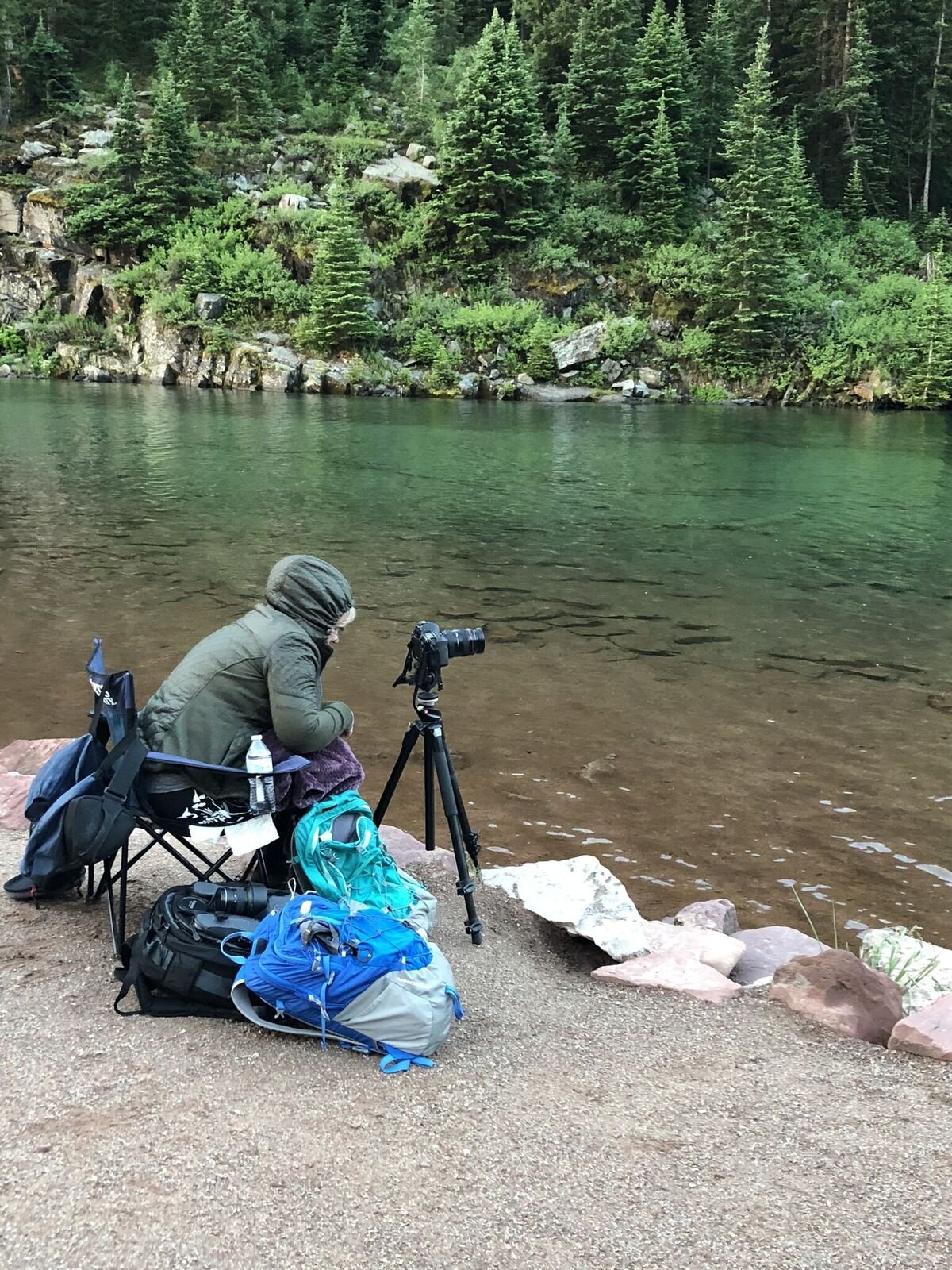 A person in outdoor clothing sits by a lake at the edge of a forested area, with camera equipment set up on a tripod, surrounded by backpacks and gear.