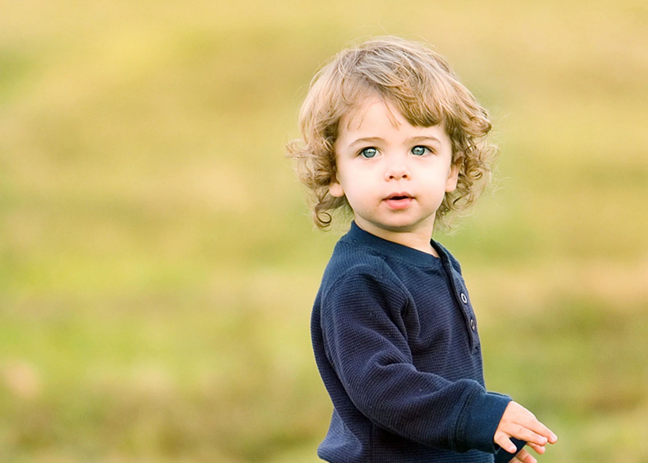 A young boy with curly blond hair and blue eyes standing outdoors with a blurred green background.