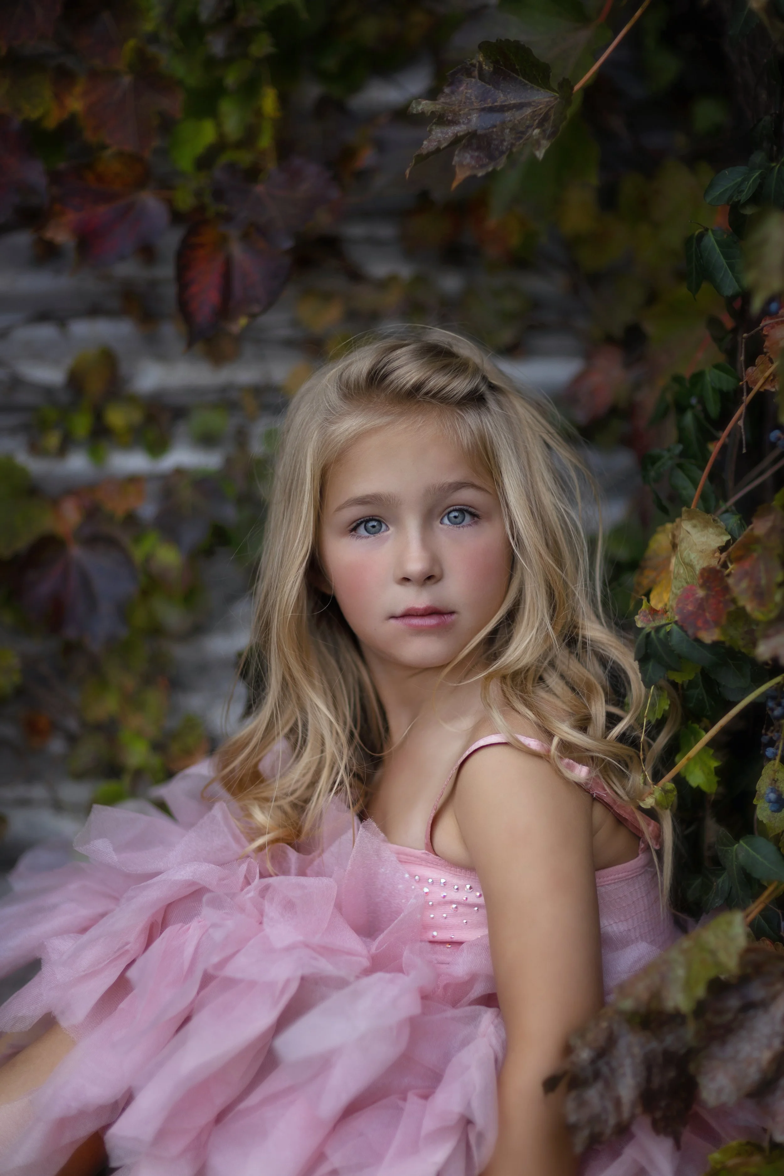 A young girl with long blonde hair and blue eyes wearing a pink dress and a pink tulle tutu in front of a background of green and brown autumn leaves.