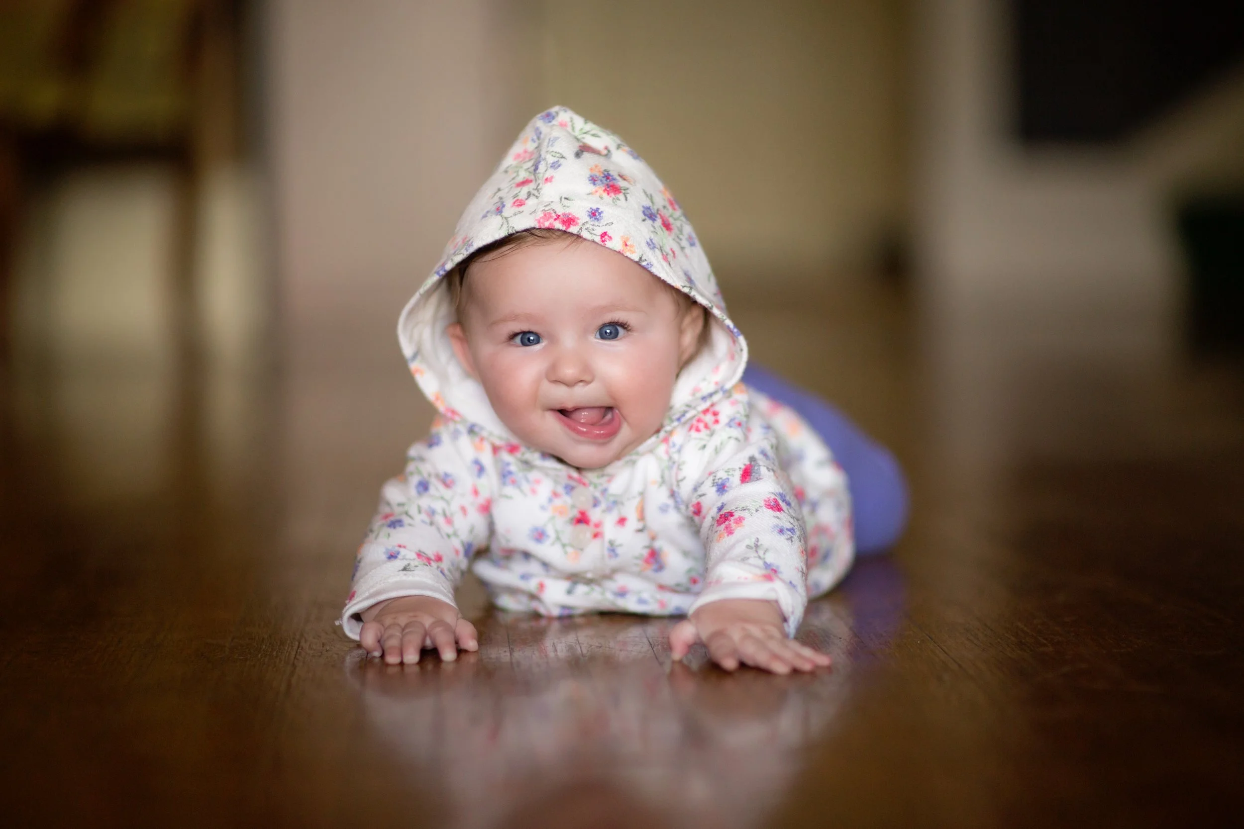A baby crawling on a wooden floor, wearing a floral hooded outfit and smiling.