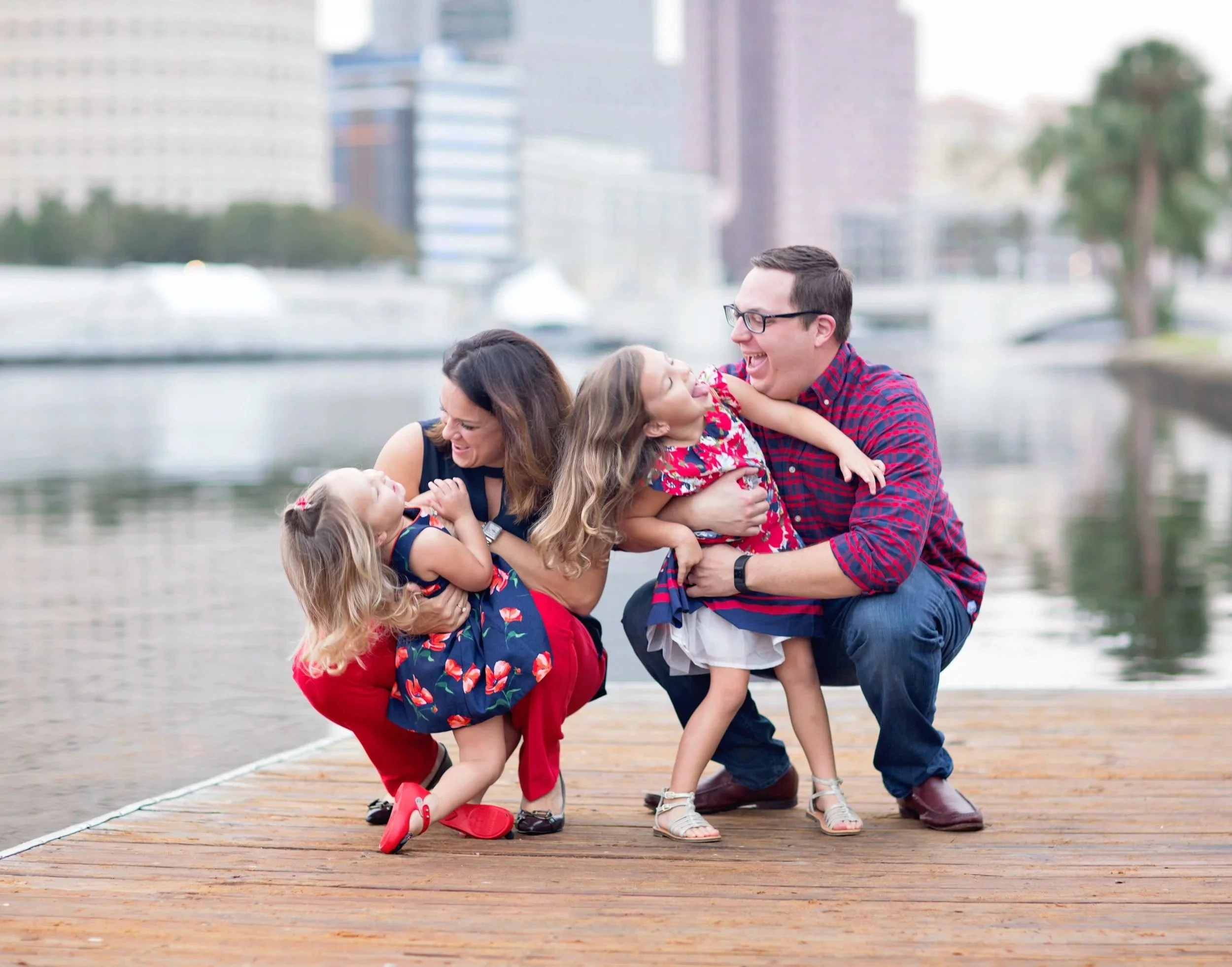 A family of four laughing and playing together on a wooden dock near a river or lake, with city buildings in the background.