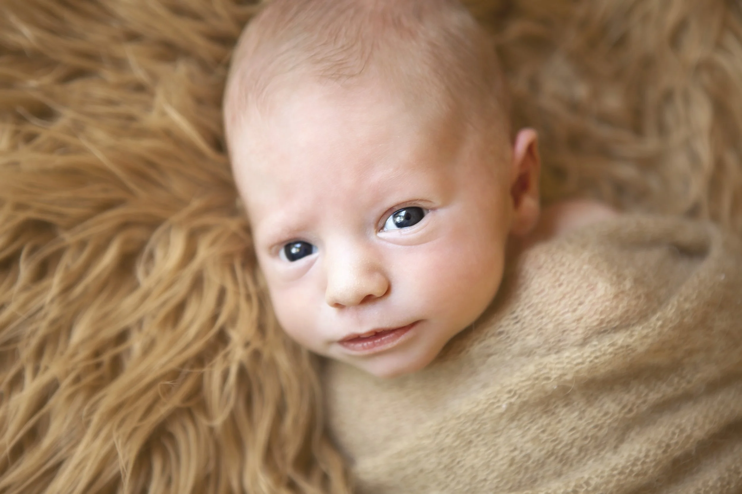 Close-up of a baby with blue eyes and light skin, lying on a soft, textured, light brown surface, with long, wavy, blonde hair surrounding the head.