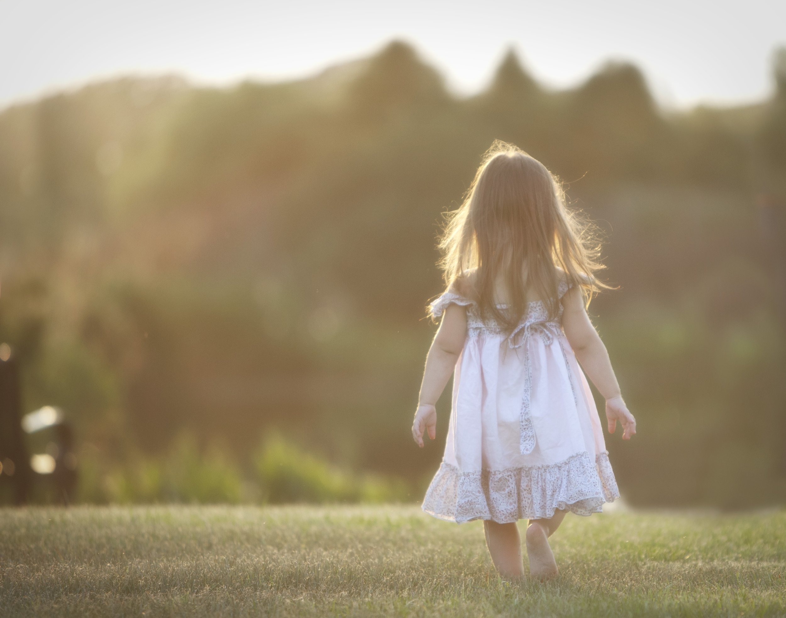 A girl in a white dress with lace details walking barefoot on grass in a park during sunset.