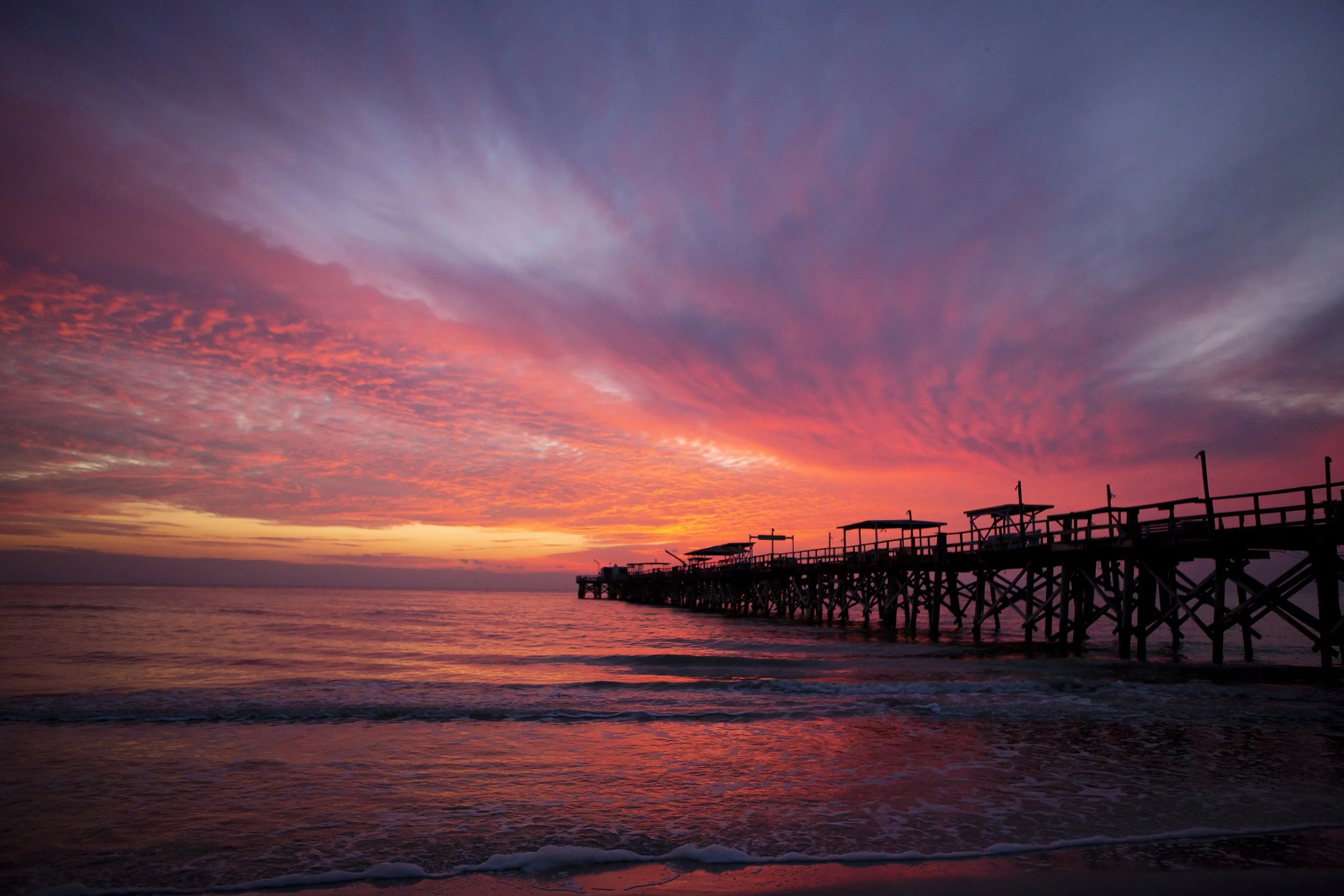 Sunset over the ocean with a wooden pier extending into the water, vibrant pink and purple clouds in the sky.