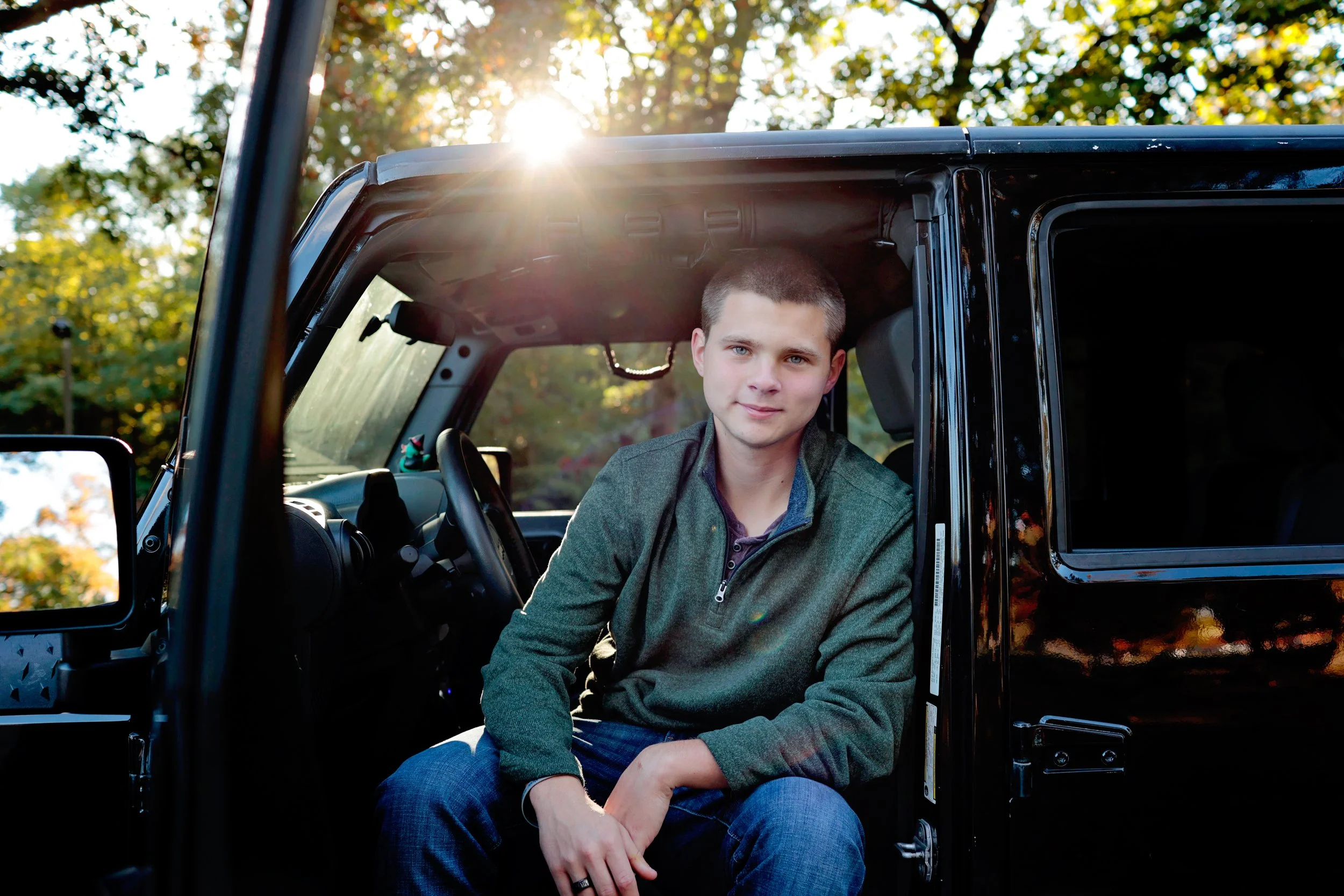 A young man wearing a green jacket and blue jeans sitting inside a black vehicle, with the door open, in a park with trees and sunlight in the background.