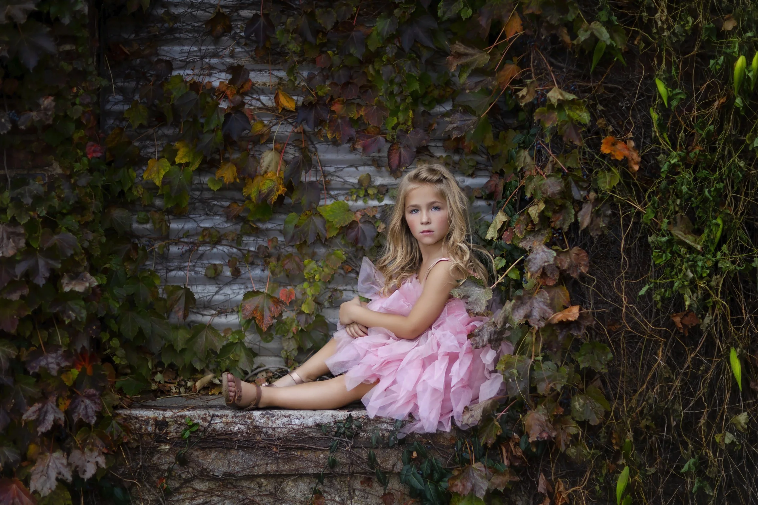 A young girl with blonde hair in a pink dress sitting on an old, rusty pipe covered with green and brown ivy leaves.