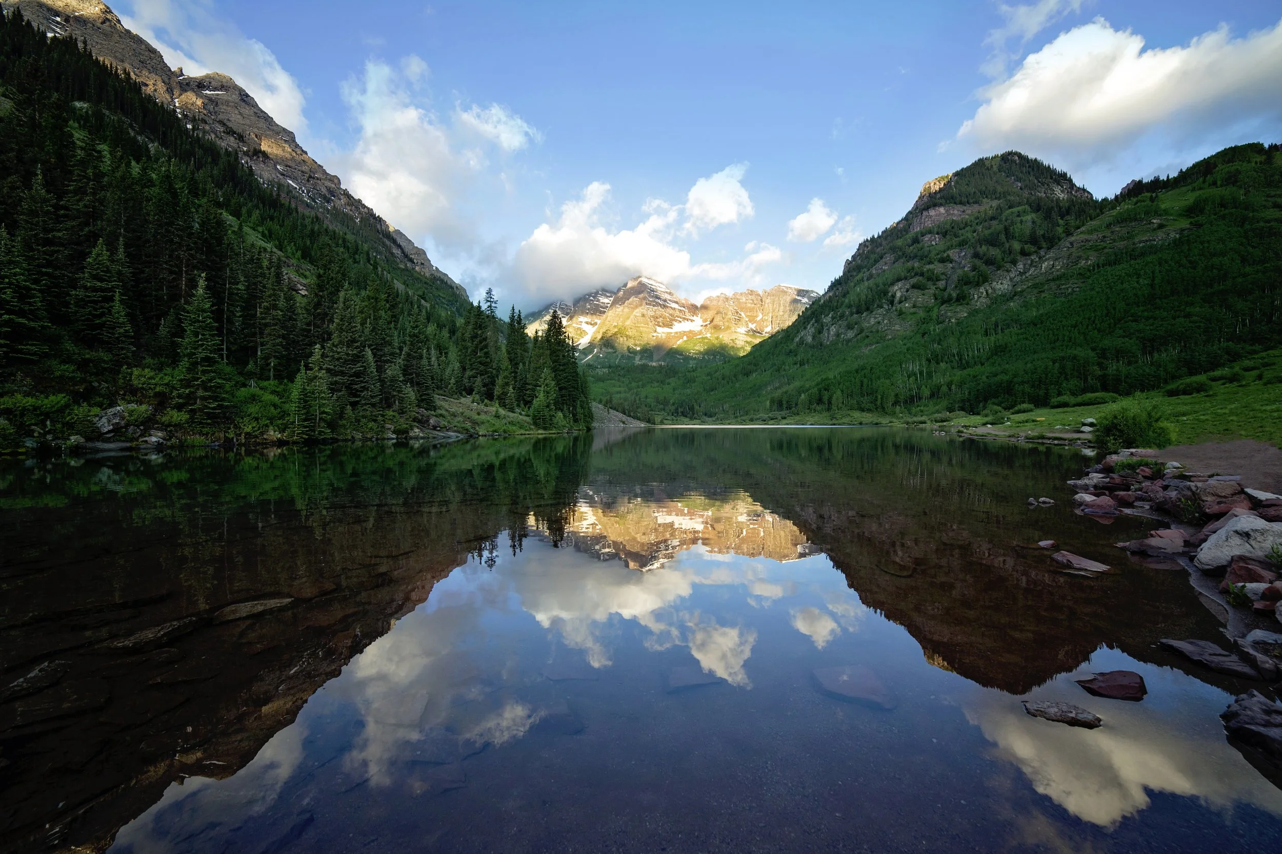 A serene mountain landscape with snow-capped peaks reflected in a calm lake surrounded by dense evergreen trees under a blue sky with white clouds.