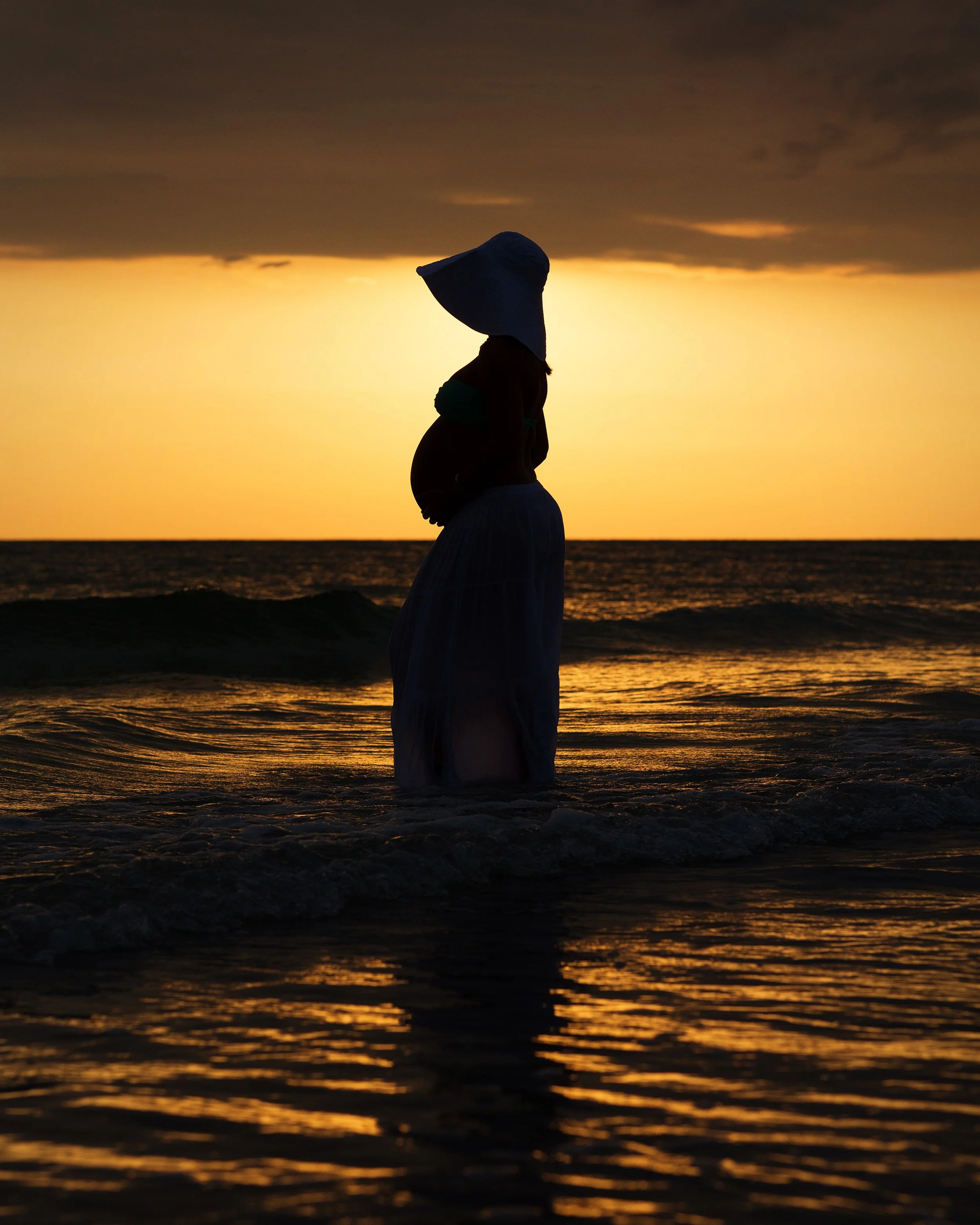 A silhouette of a person wearing a wide-brimmed hat at the beach during sunset, with dark clouds above and the ocean reflecting the orange and yellow hues of the sky.