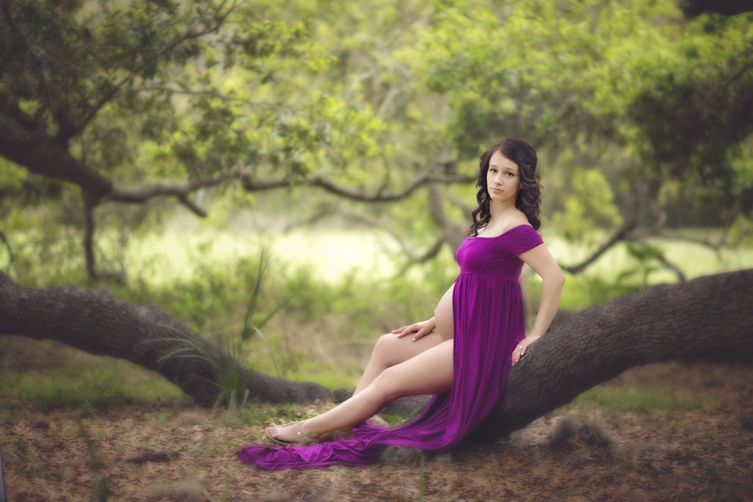 A pregnant woman in a purple dress sitting on a tree branch outdoors in a forested area.