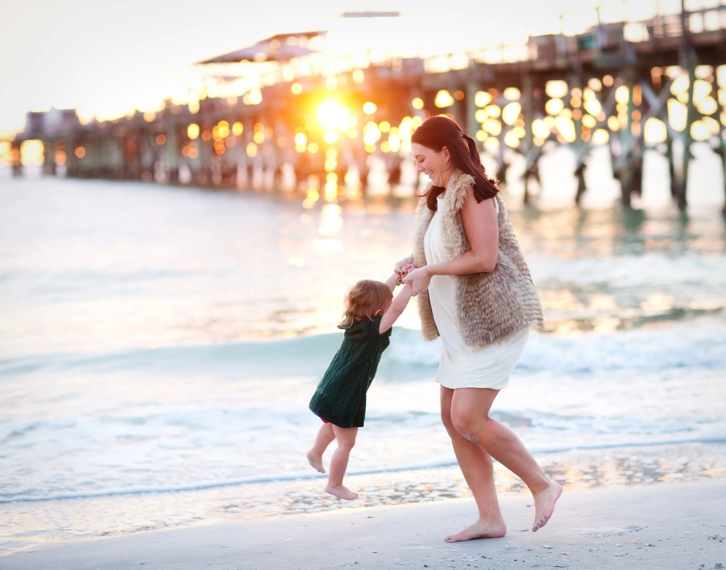 A woman and two children playing near the ocean at sunset, with pier and bright sunlight in the background.