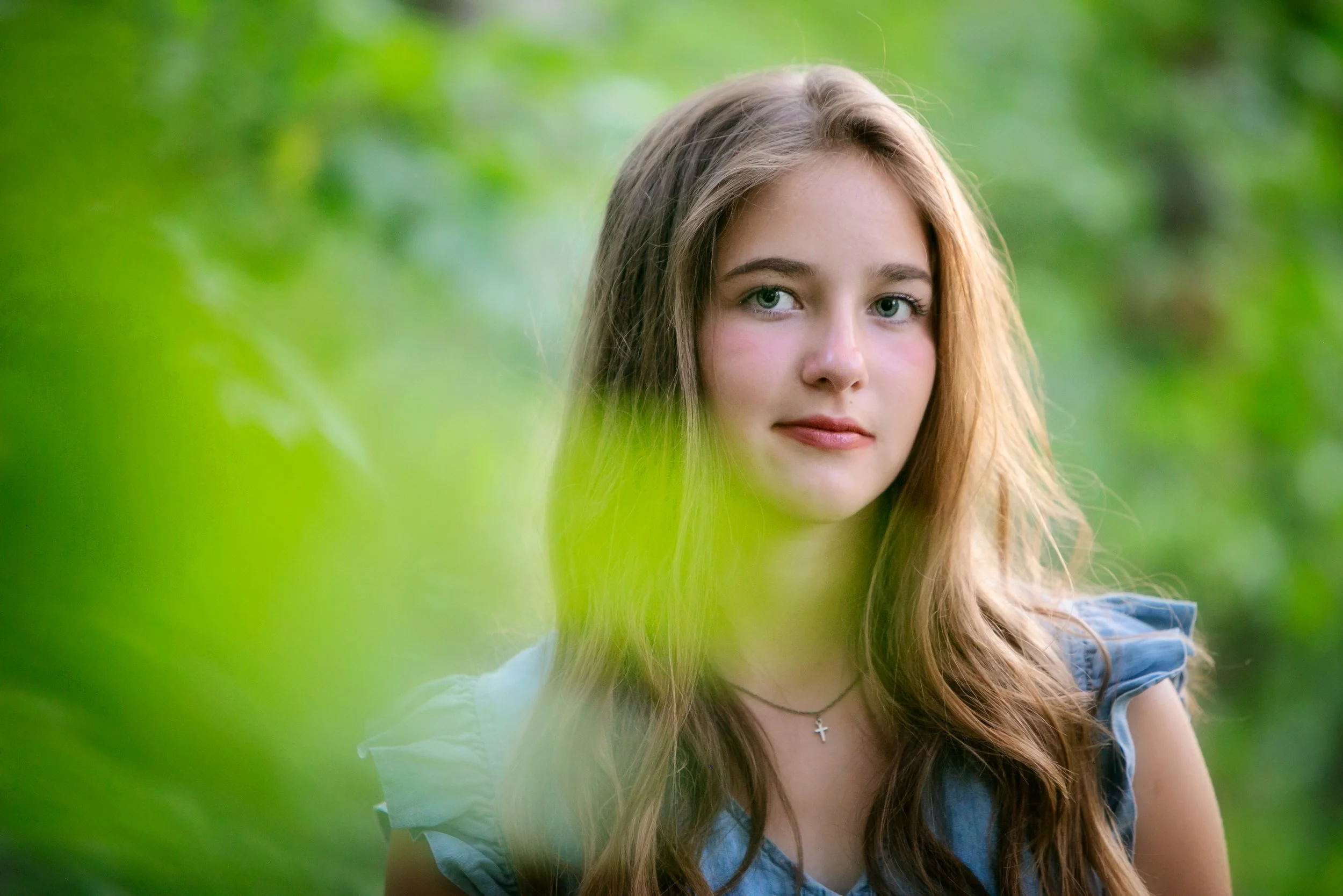 A young girl with long brown hair, wearing a blue top and a cross necklace, standing outdoors with a blurred green background.