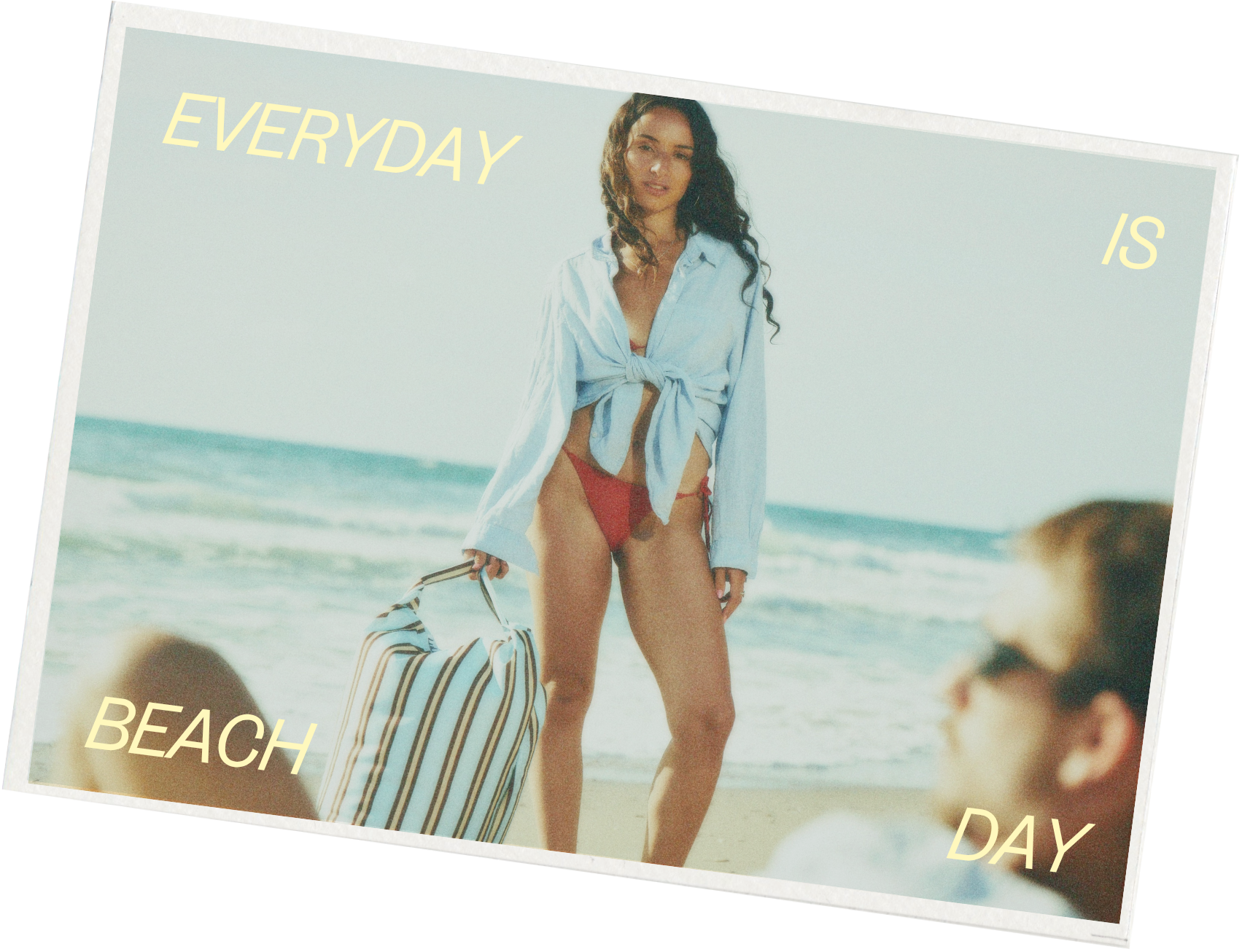 A woman in a light blue tied shirt and red bikini bottom standing on the beach with the ocean in the background, holding a striped bag, with the words "Everyday is beach day" written on the image.