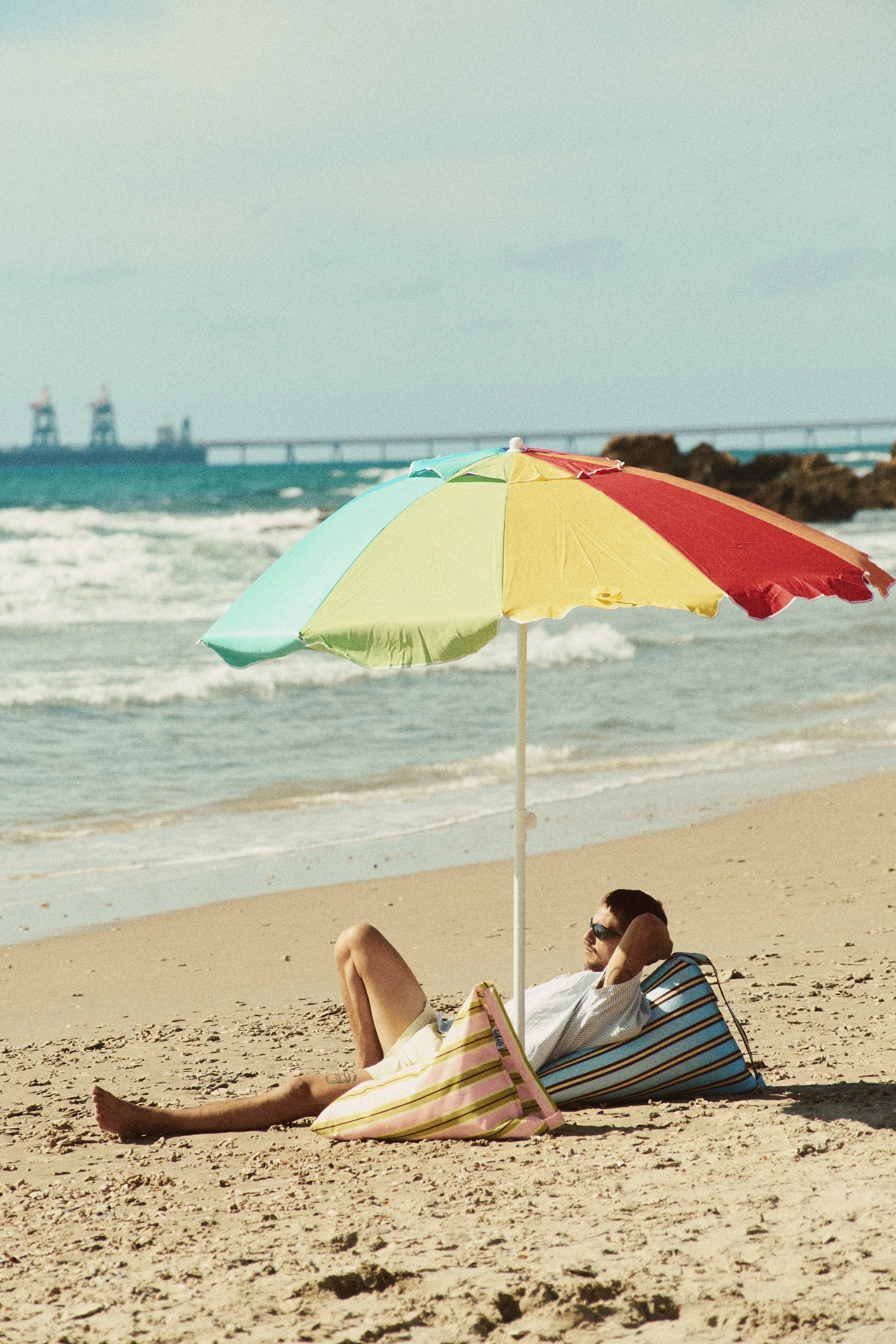 A man relaxing on a striped beach chair under a colorful umbrella on a sandy beach with waves and rocks, with a bridge and two oil rigs in the background.