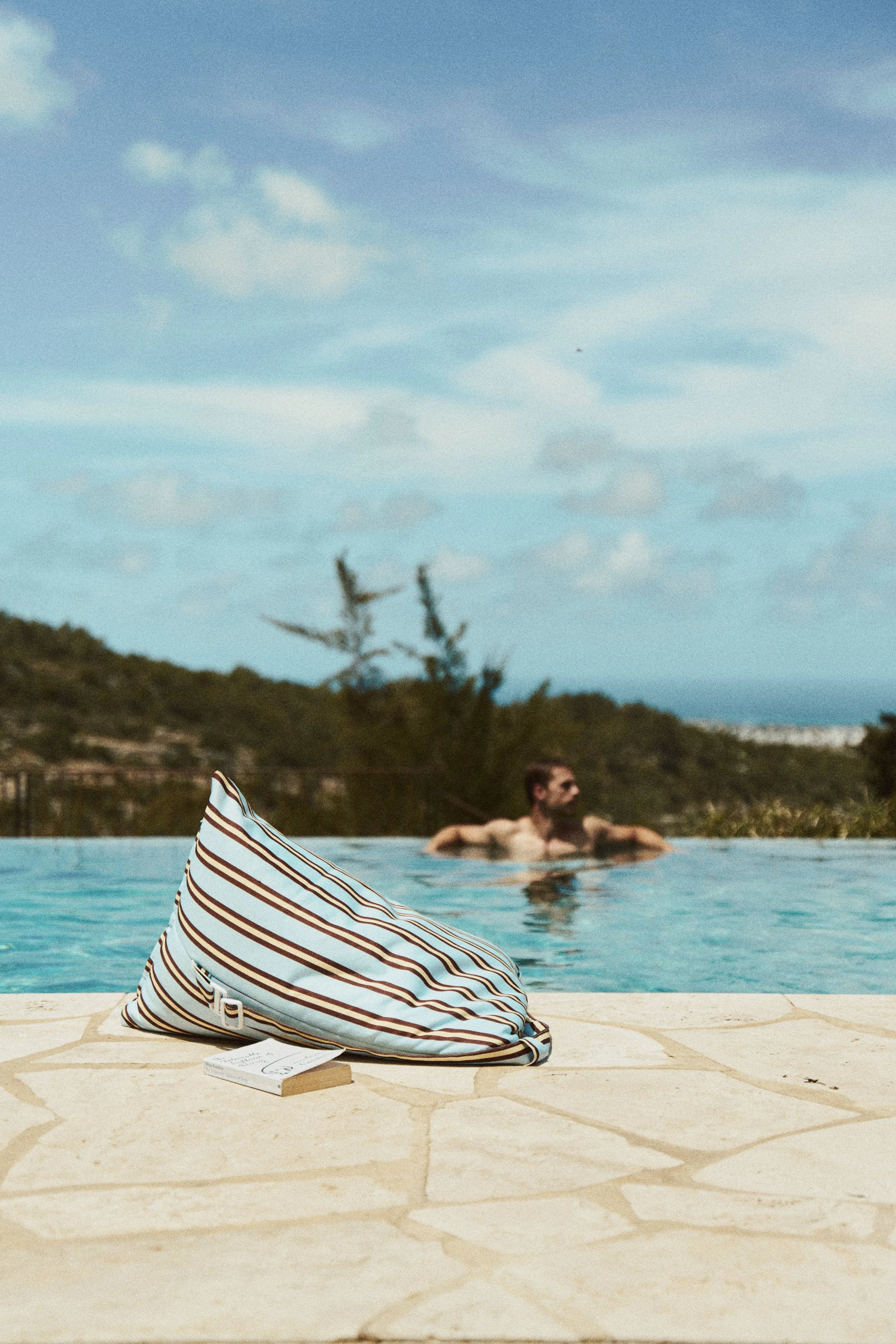 A striped bag and a book with a pen on top are placed on a stone surface by a swimming pool. A person is swimming in the pool in the background with a scenic mountain and sky view.