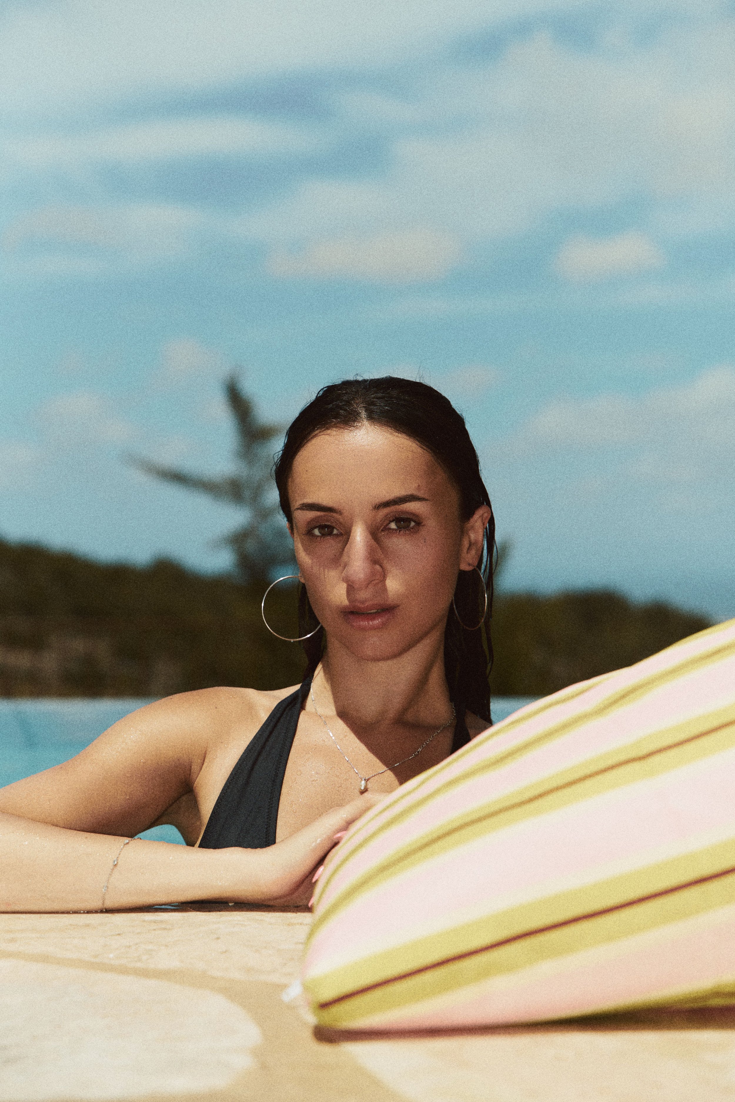 A woman in a black swimsuit lying on a beach with a blue sky and clouds in the background, and striped yellow and pink items in the foreground.