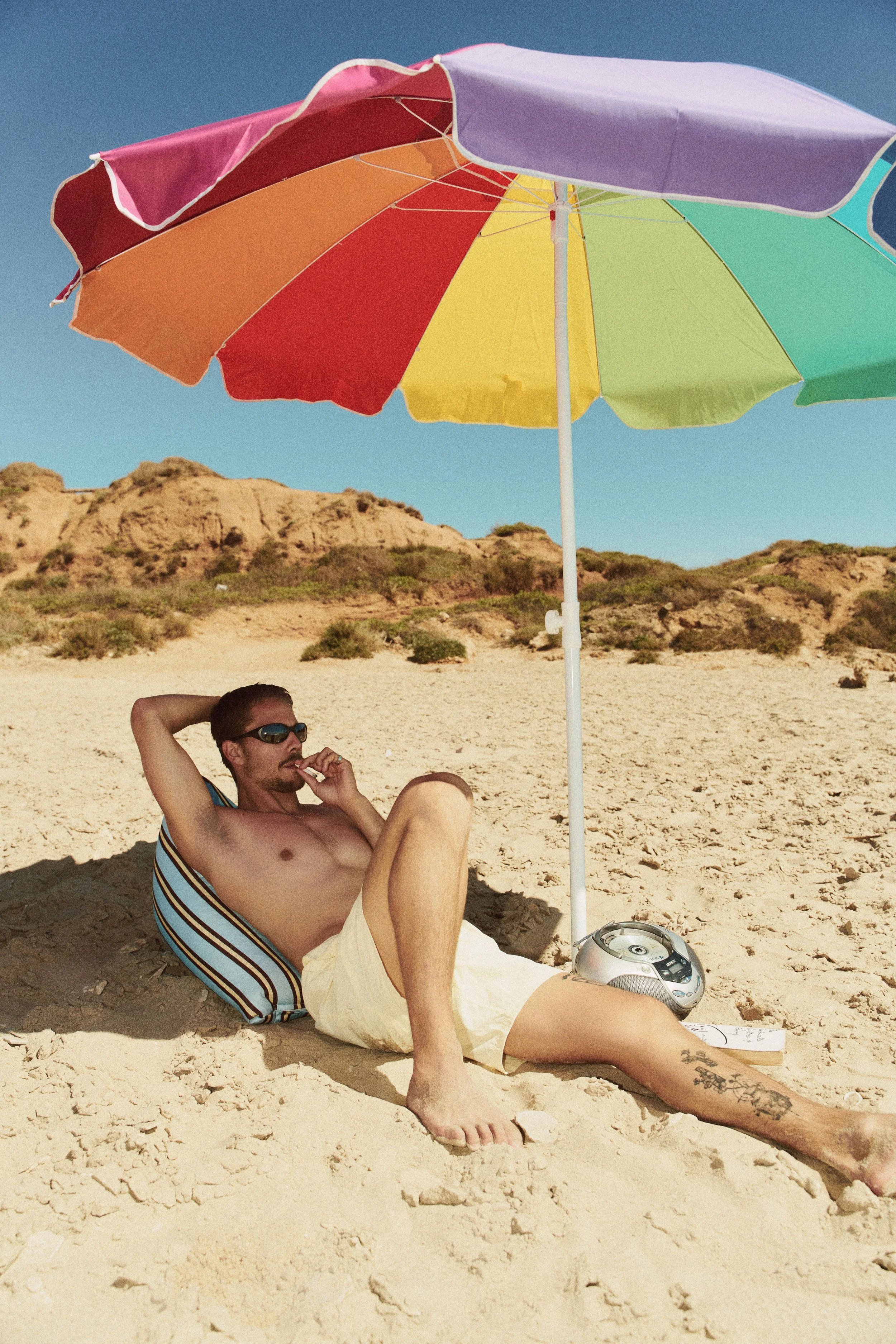 A man relaxing on a sandy beach under a colorful umbrella, wearing sunglasses and striped sleeveless shirt, with a portable stereo and a notebook nearby.