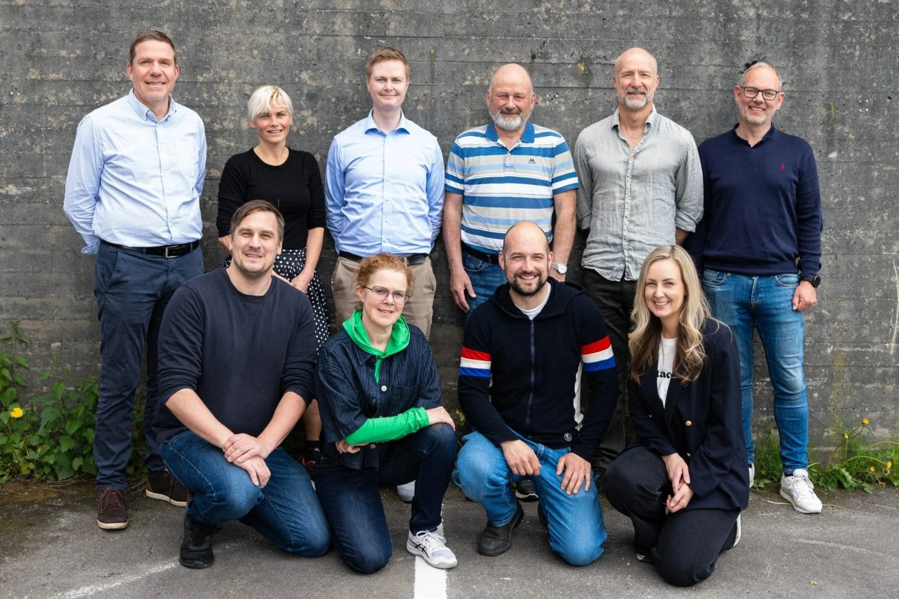 Et gruppe mennesker poserer for et bilde foran en murvegg, noen sitter og noen står.