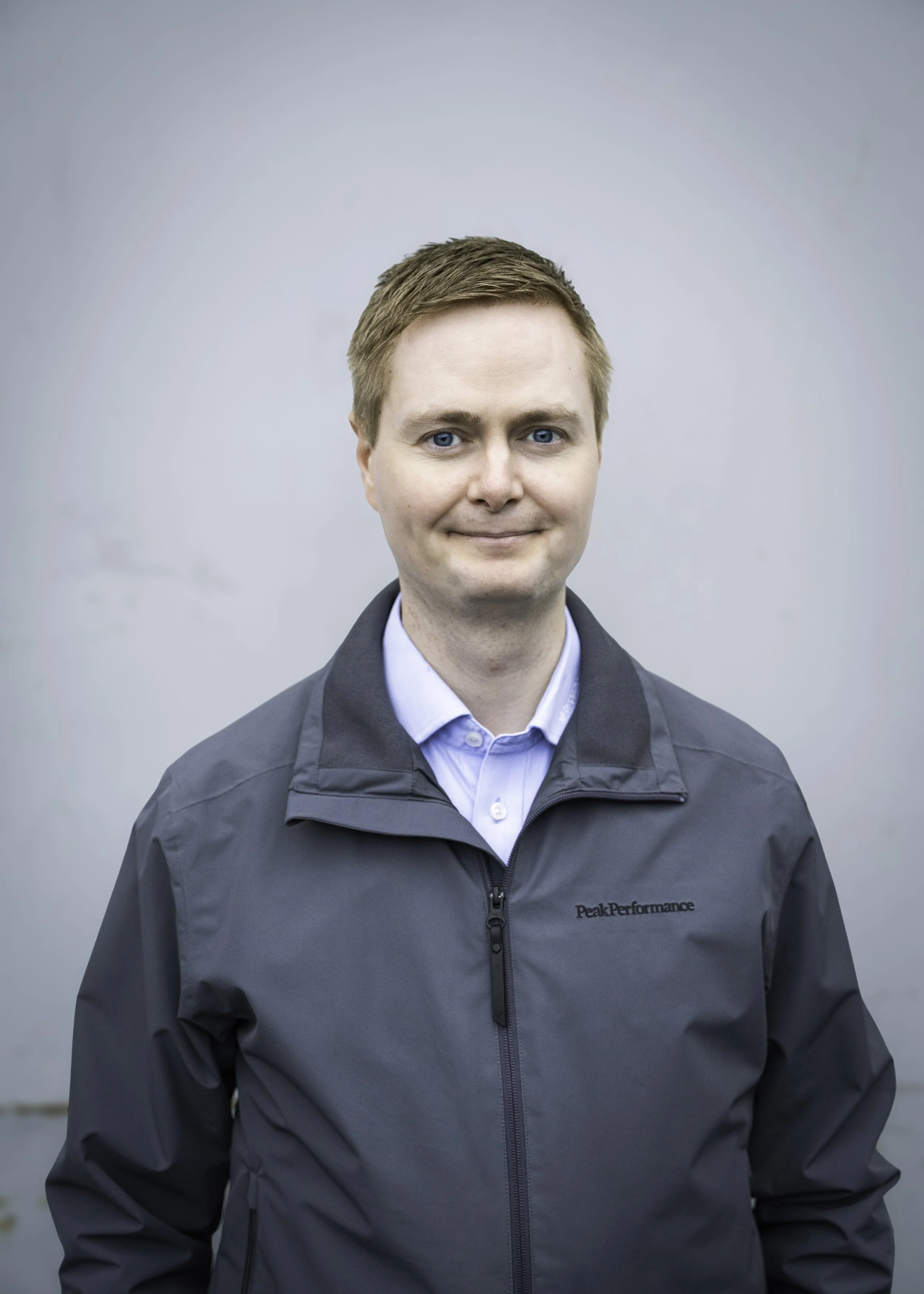 Portrett av en mann med lyse, kort hår, kledd i en grå Peak Performance jakke og lys blå skjorte, mot en grå, ustrukturert bakgrunn.