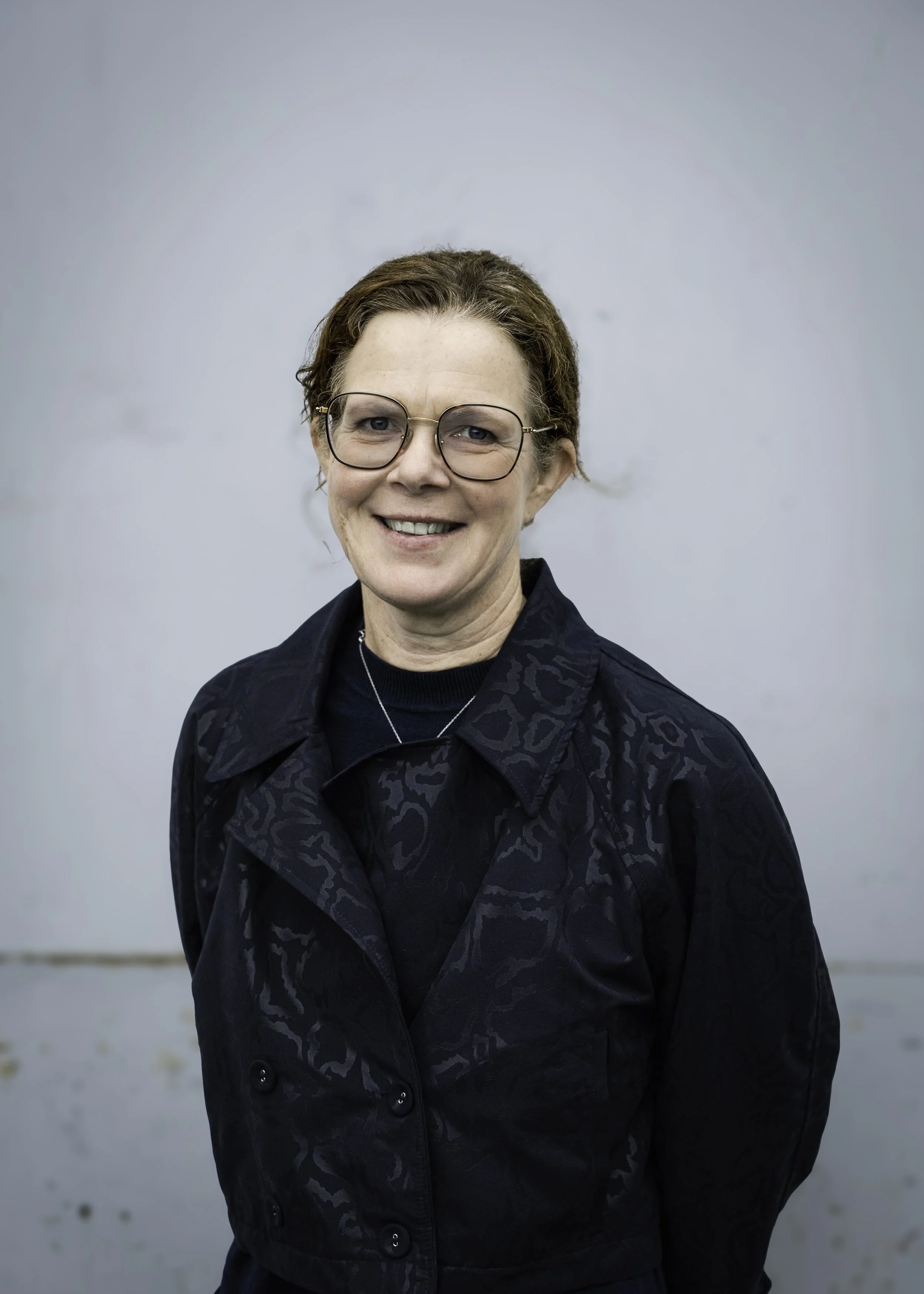 Portrettbilde av en kvinne med brunt, krøllet hår og store, runde briller, smilende mot kameraet, iført en mørk, mønstret jakke, stående foran en enkel, lys bakgrunn.