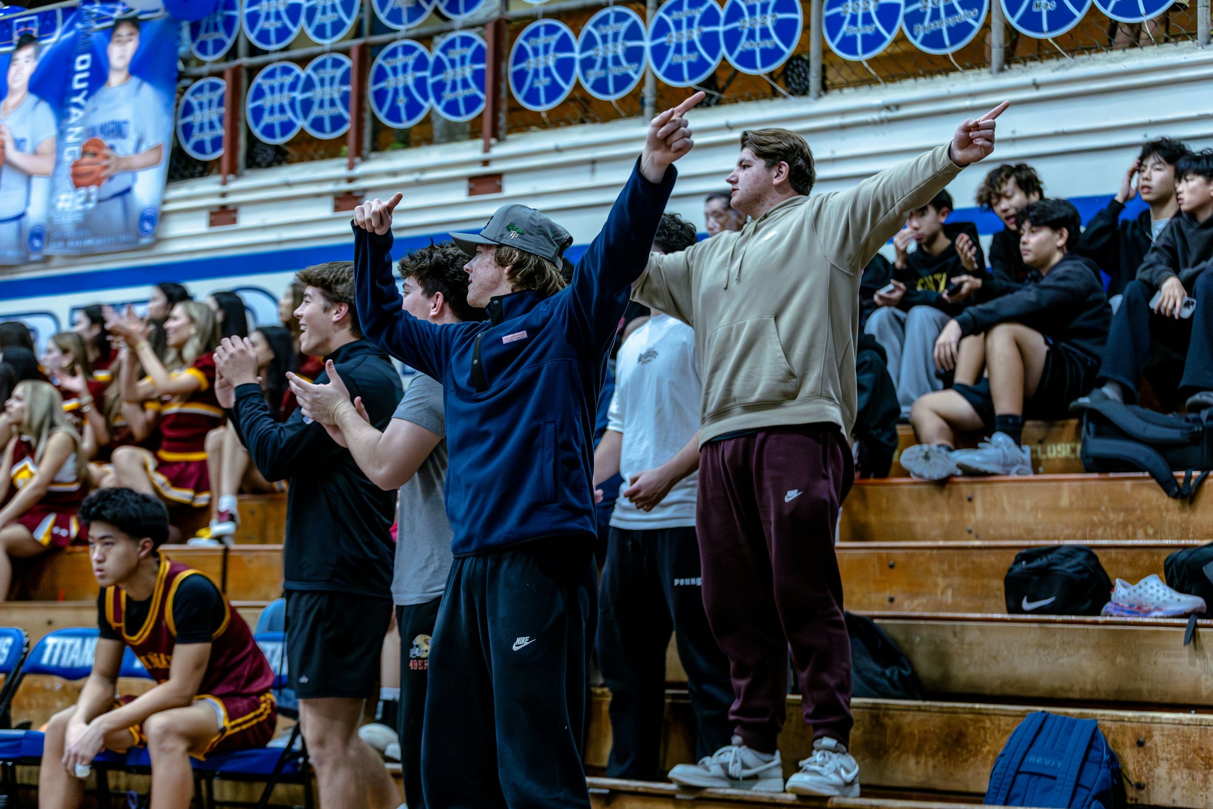 Spectators at an indoor sports event, seated on wooden bleachers, with two young men standing and pointing, surrounded by other young people and cheerleaders in the background.