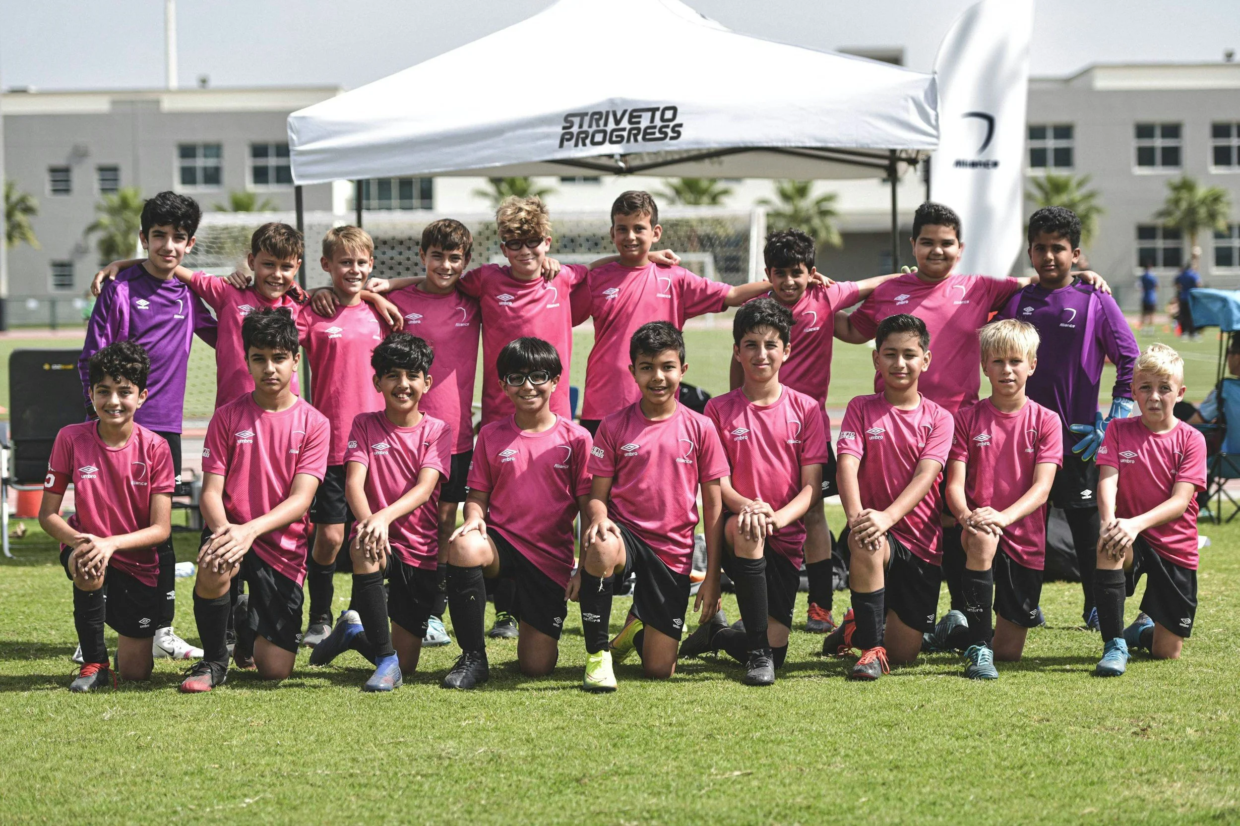 A group of young boys in pink and purple soccer uniforms posing together on a grassy field, some standing and some kneeling, with two adults behind them under a white canopy. Buildings and trees are visible in the background.