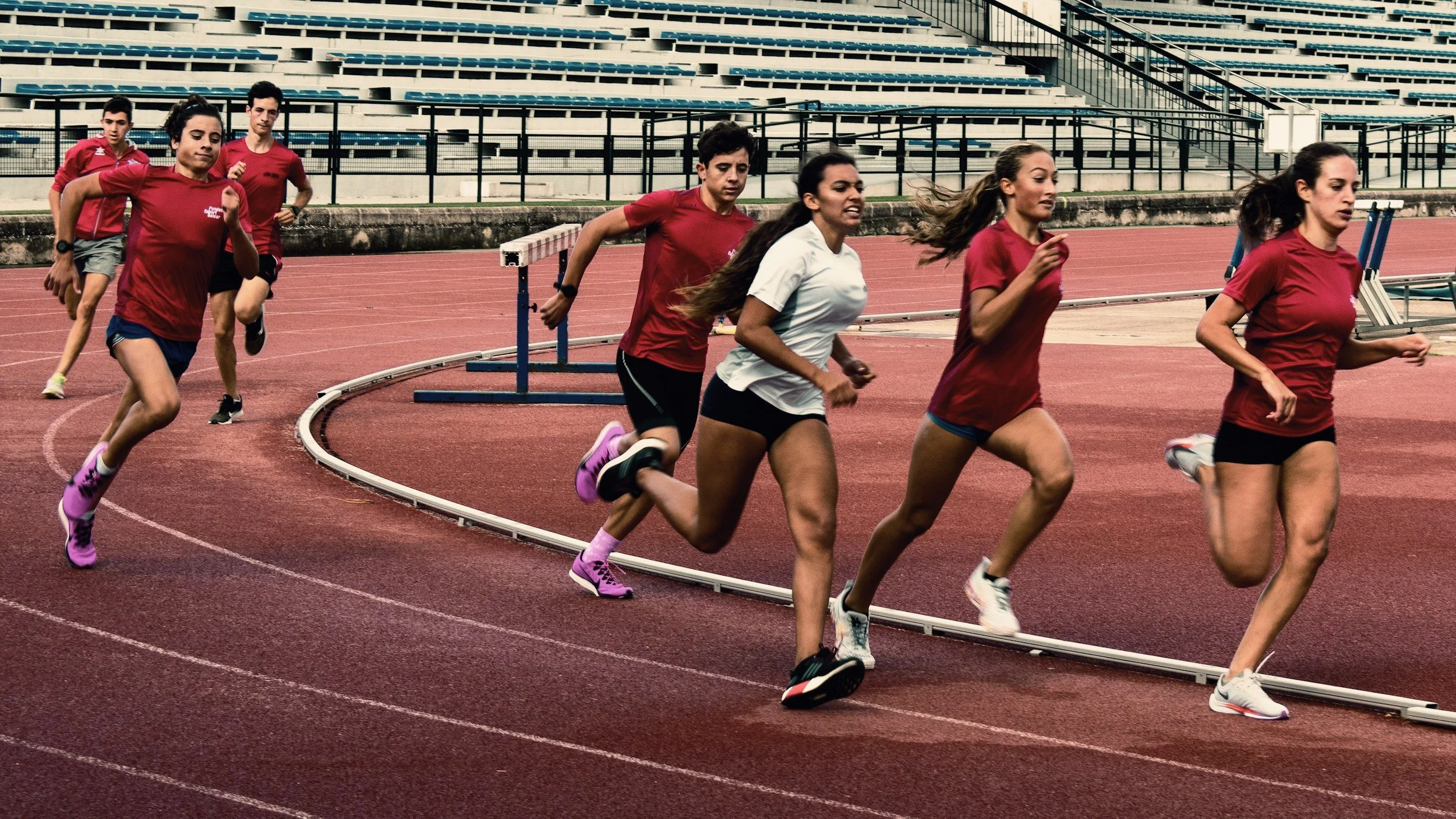 A group of women and girls running on a curved red track in a stadium, wearing athletic clothing.