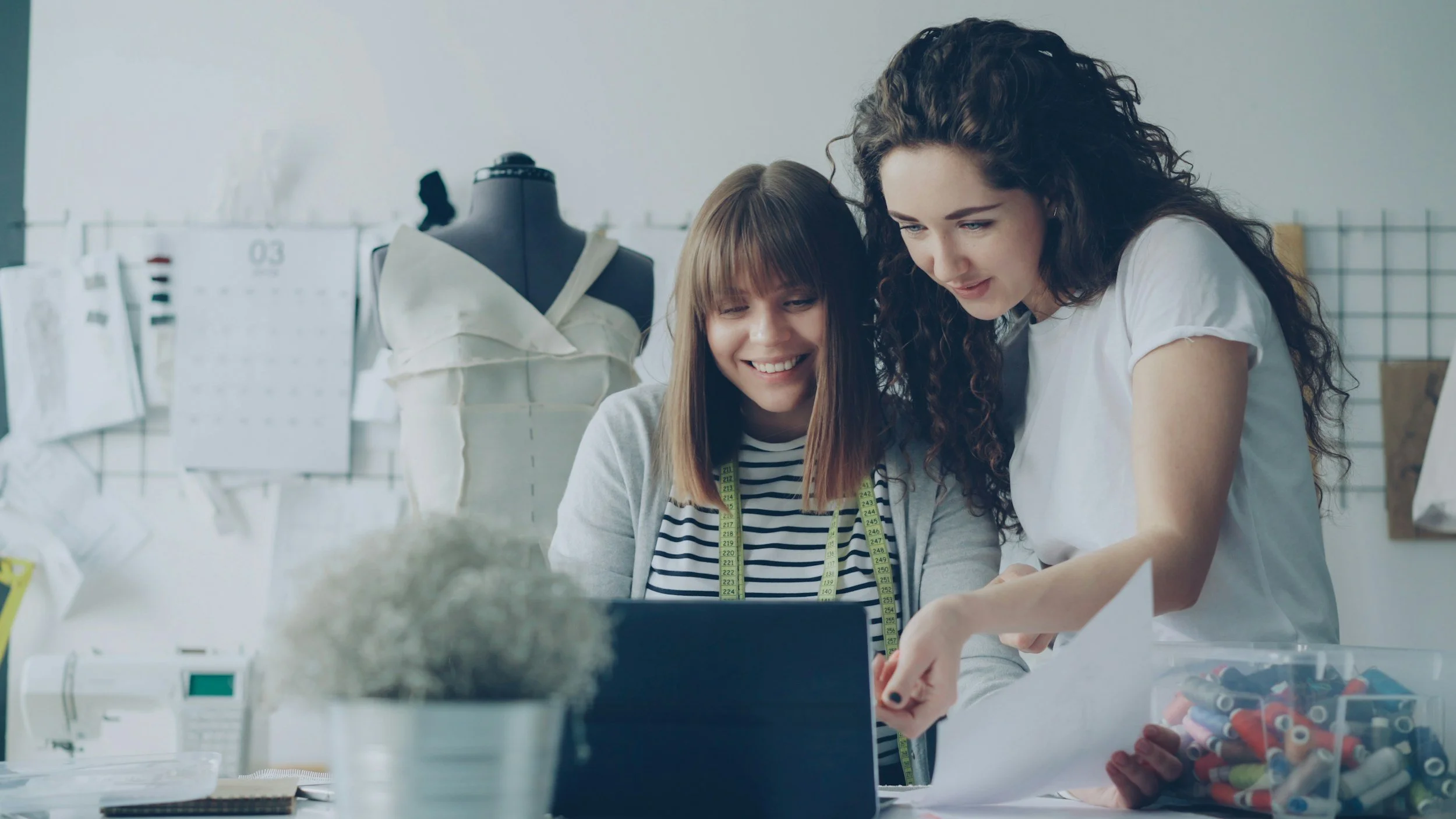Two women working together at a sewing or fashion design workspace, with fabric and sewing tools, in a bright studio.