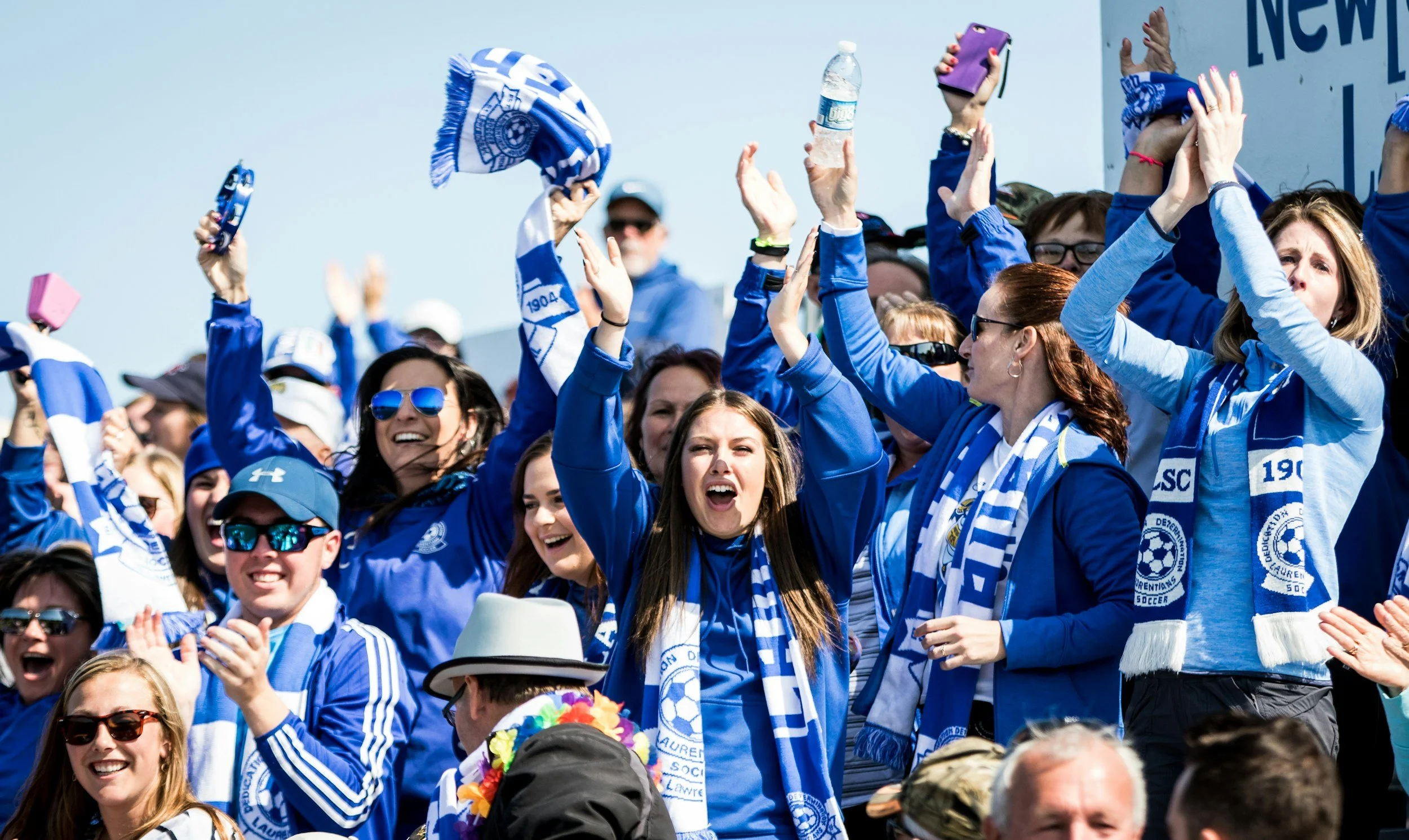 A crowd of people wearing blue and white sports apparel and scarves, celebrating at an outdoor event, with some holding hats and others waving their hands.