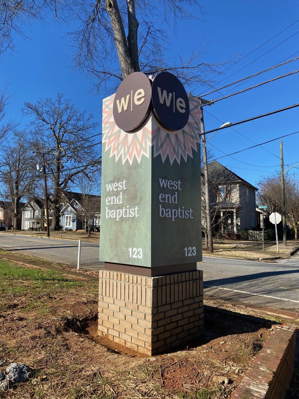 Sign on a brick pedestal reading "West End Baptist" at 123 with two circular signs above displaying "we" on a gray and black background, respectively, and a decorative pattern behind.