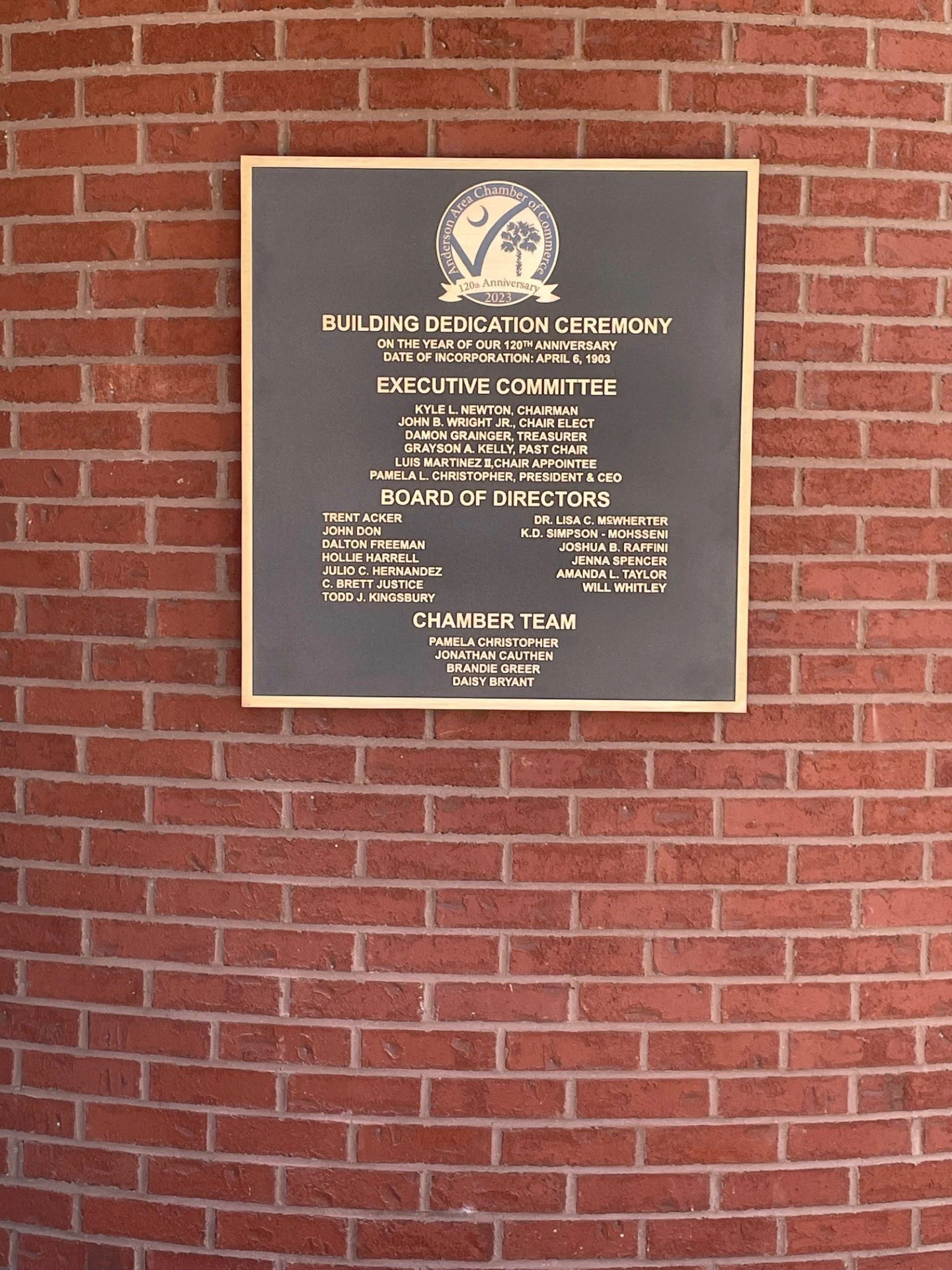 A framed plaque mounted on a red brick wall, displaying information about a building dedication ceremony for the Anderson Area Chamber of Commerce's 120th anniversary in 2023.