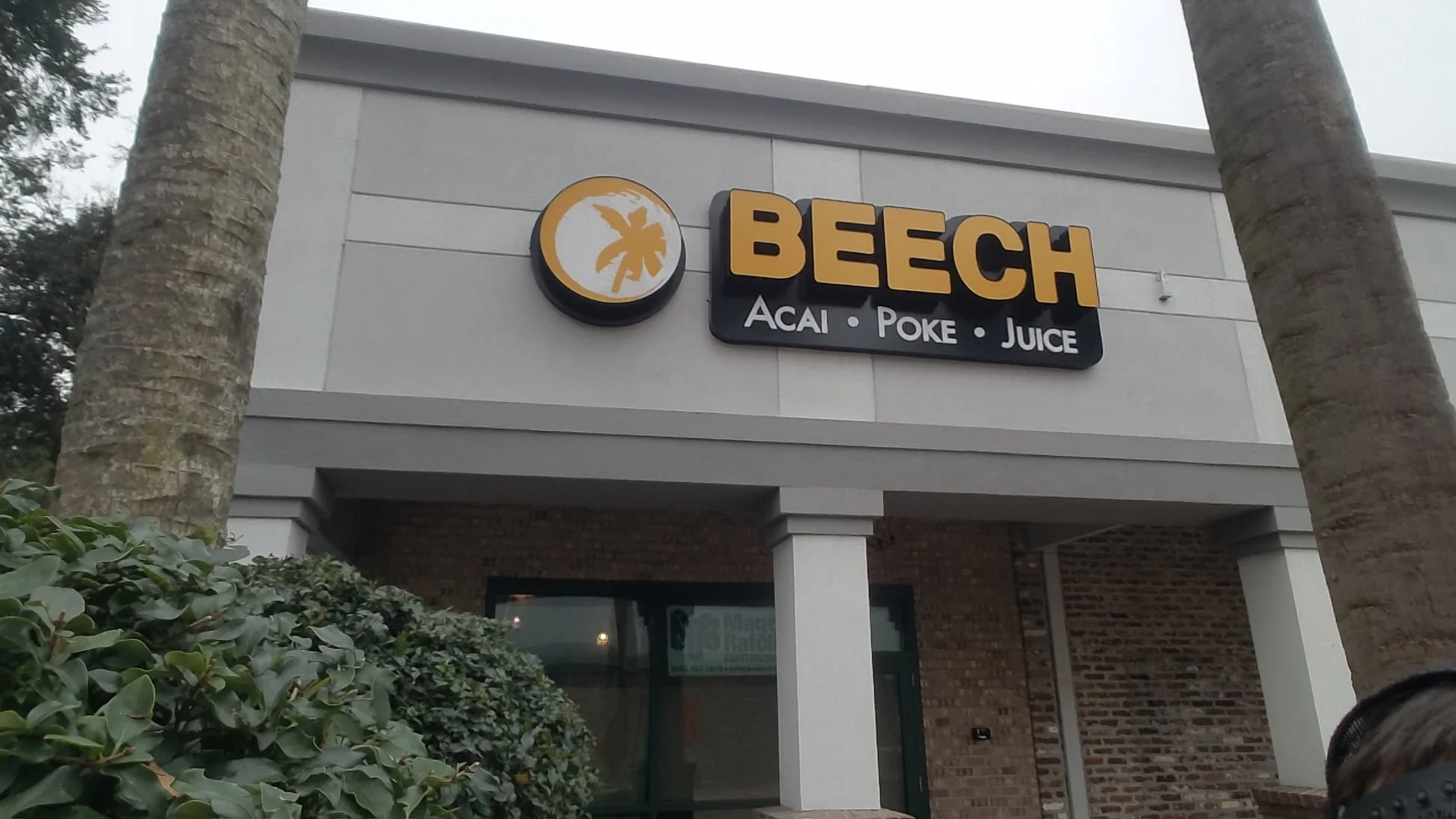 Exterior of a store named Beech, selling acai, poke, and juice, with palm trees and greenery in front.