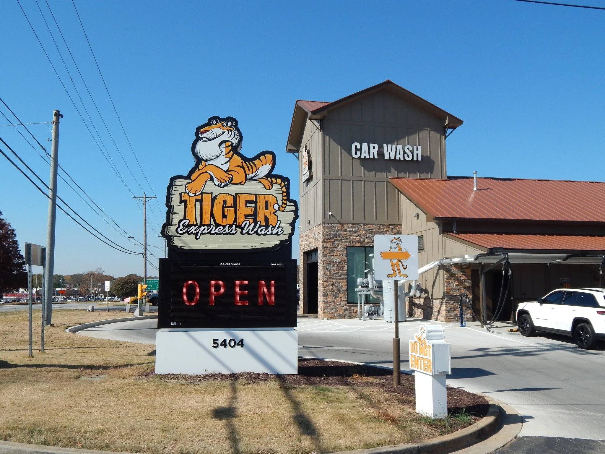 Sign for Tiger Express Wash car wash with a cartoon tiger mascot sitting on a wooden sign, and a building with a "Car Wash" sign on it.