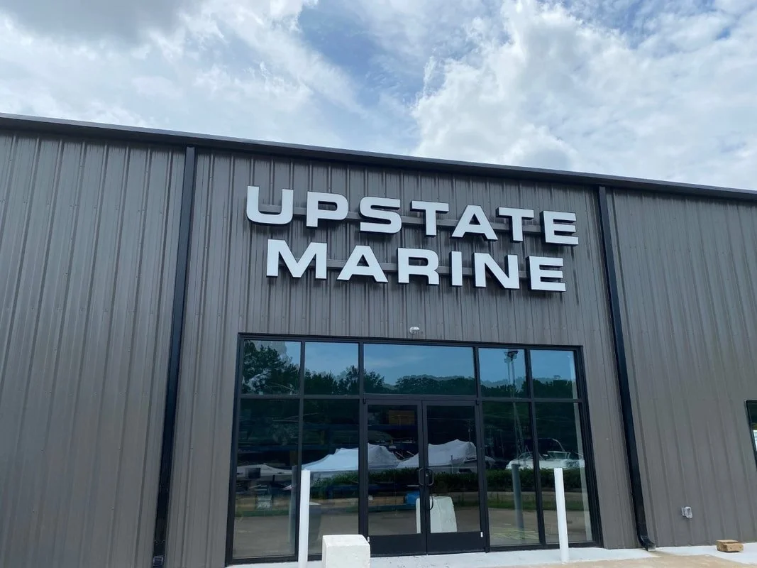 The storefront of Upstate Marine with large black metal letters spelling out its name, gray exterior walls, glass doors, and windows reflecting the outdoors, with a partly cloudy sky above.