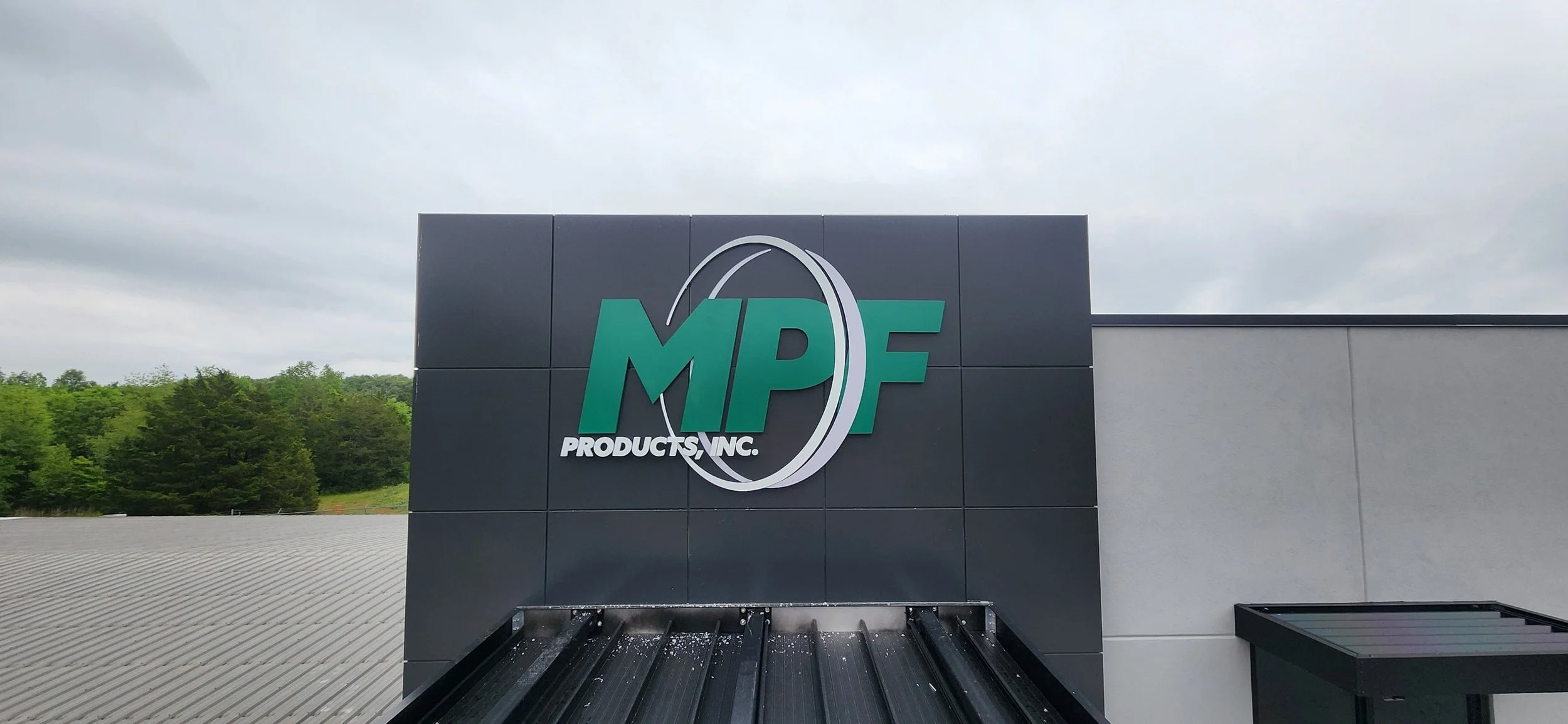 Exterior sign of MPF Products Inc. on a building, with a cloudy sky and green trees in the background.