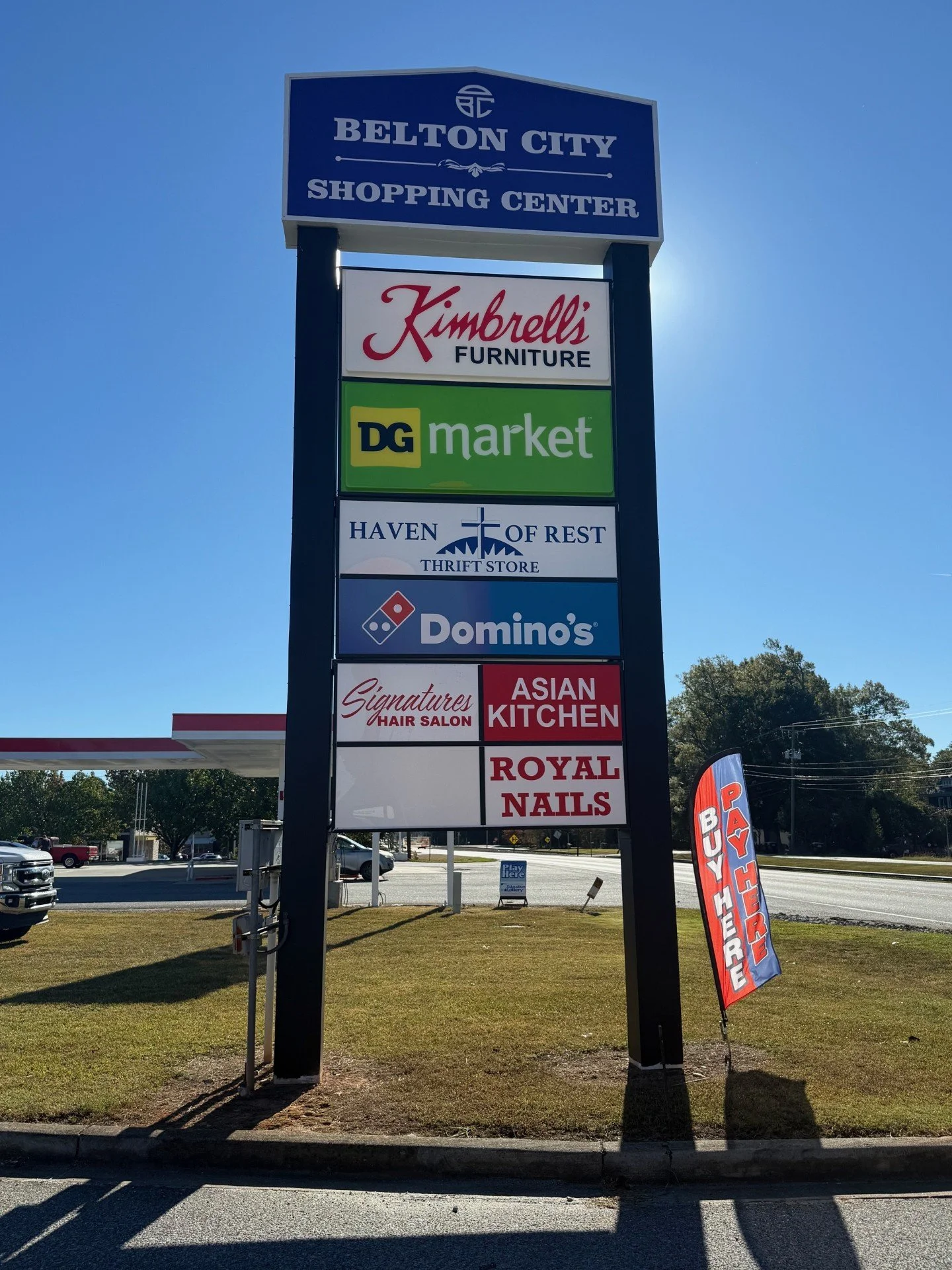 A tall sign listing several businesses at Belton City Shopping Center, including Kimberly's Furniture, DG Market, Haven of Rest Thrift Store, Domino's, Signatures Hair Salon, Asian Kitchen, and Royal Nails. There is a red and blue vertical flag with 
