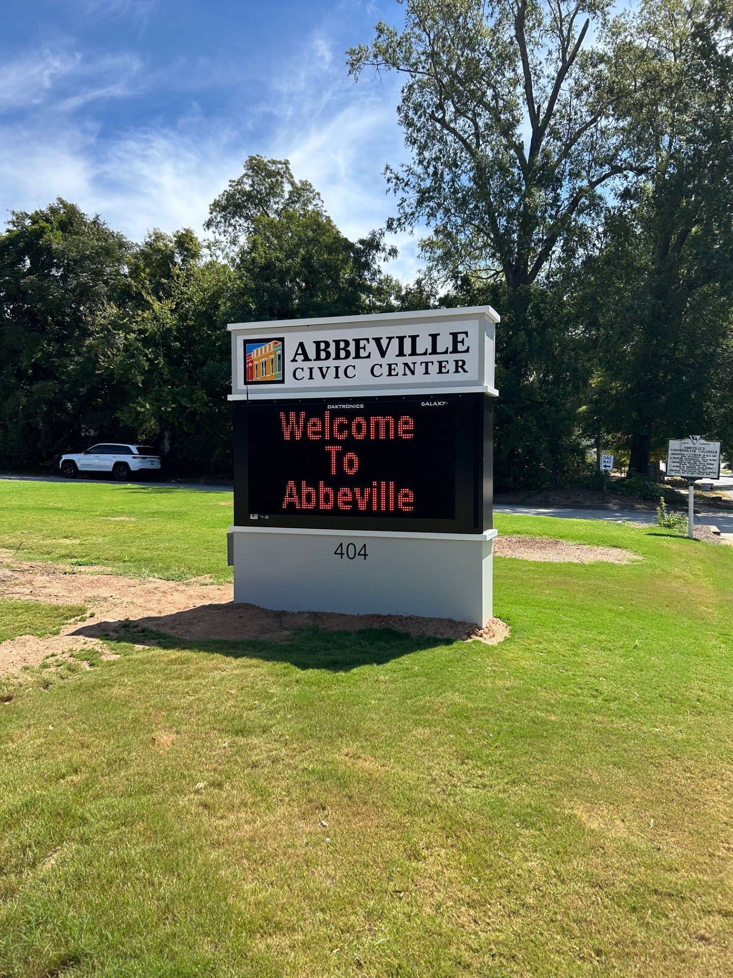 Sign at Abbeville Civic Center reading "Welcome to Abbeville" with a colorful logo, situated on a well-kept lawn with trees and a clear blue sky in the background.