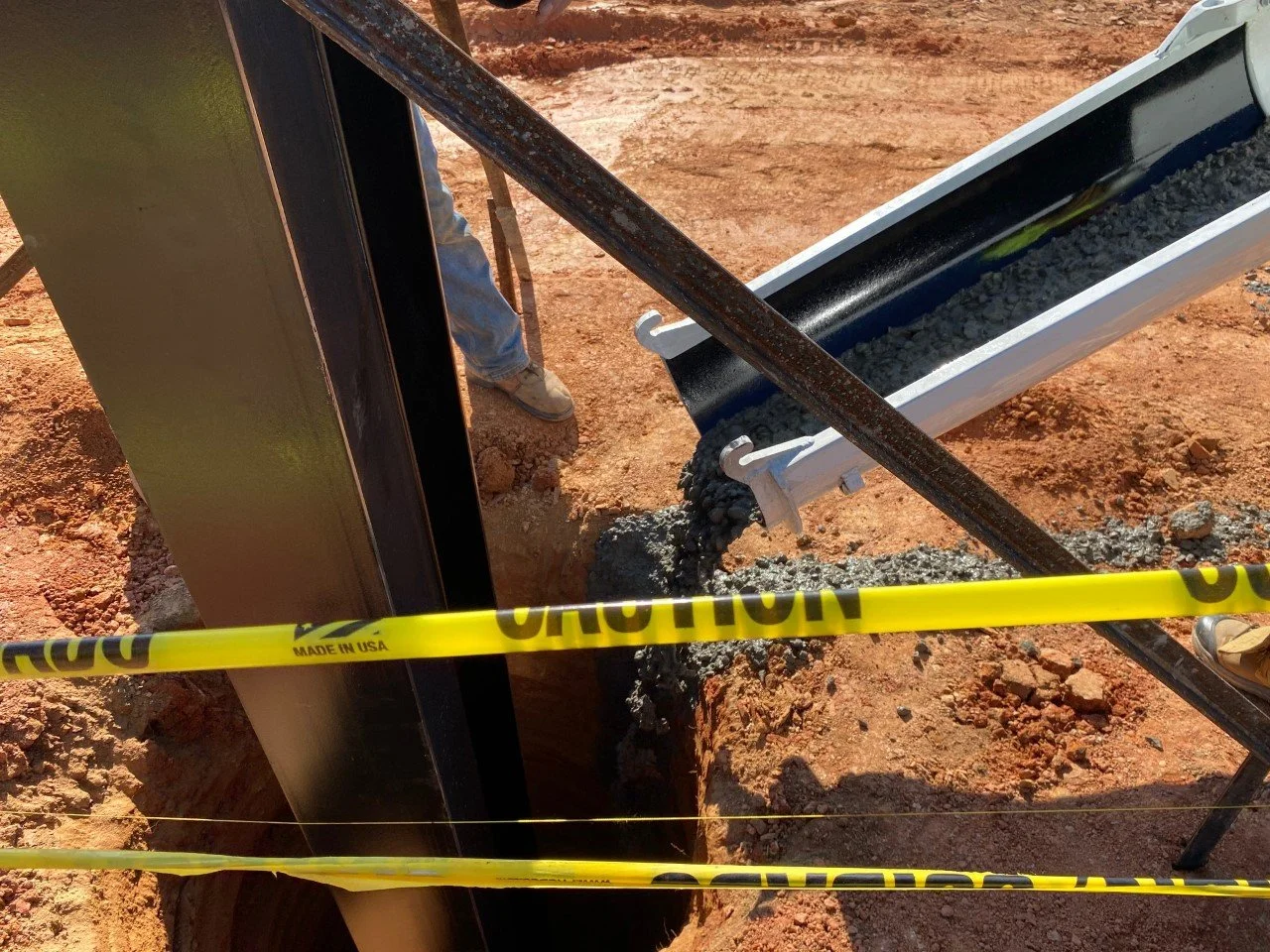 Construction site with a large black vertical structure, a conveyor dispensing gravel or small stones into a trench, with yellow caution tape in the foreground and two workers' legs visible.