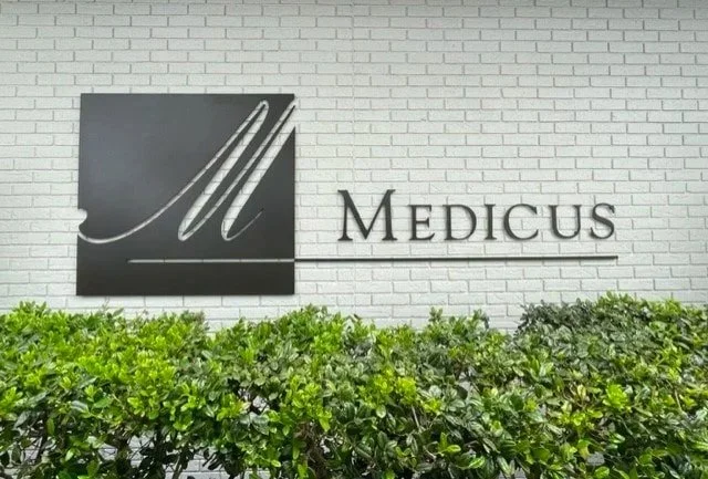 Sign on a white brick wall displaying a black square with a silver 'M' logo and the word 'MEDICUS' in black letters underneath.
