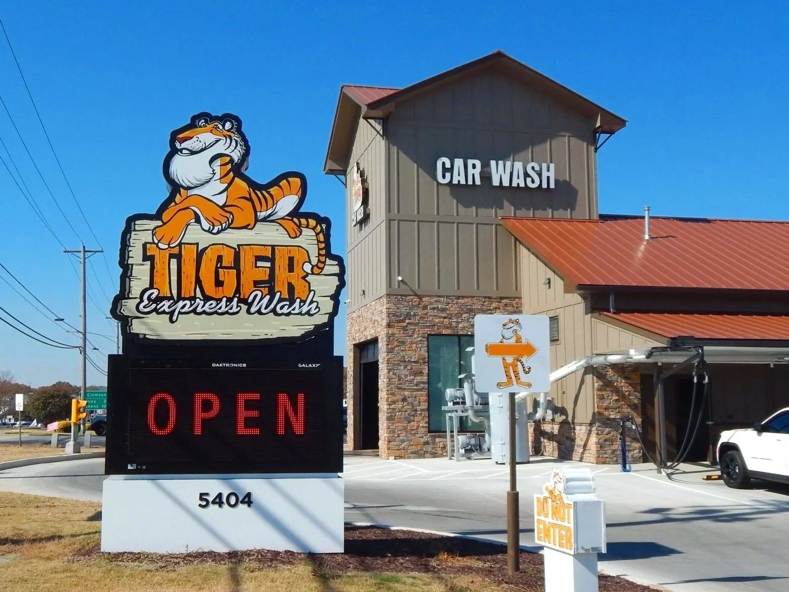 A sign for Tiger Express Wash with a cartoon tiger lying on a wooden sign, and the words 'Tiger Express Wash' in bold, stylized letters. The sign is outside a building with a red metal roof, and additional signs indicating a car wash and drive-in ent