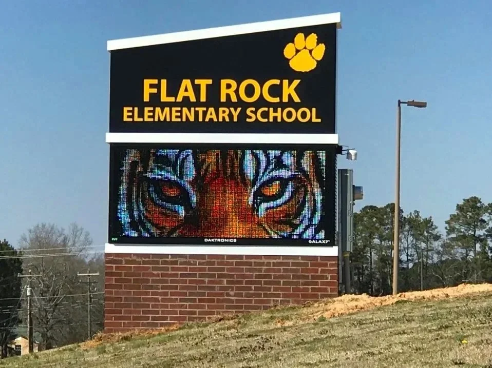 Sign for Flat Rock Elementary School with a yellow paw print and a digital display showing a close-up image of a tiger's face.