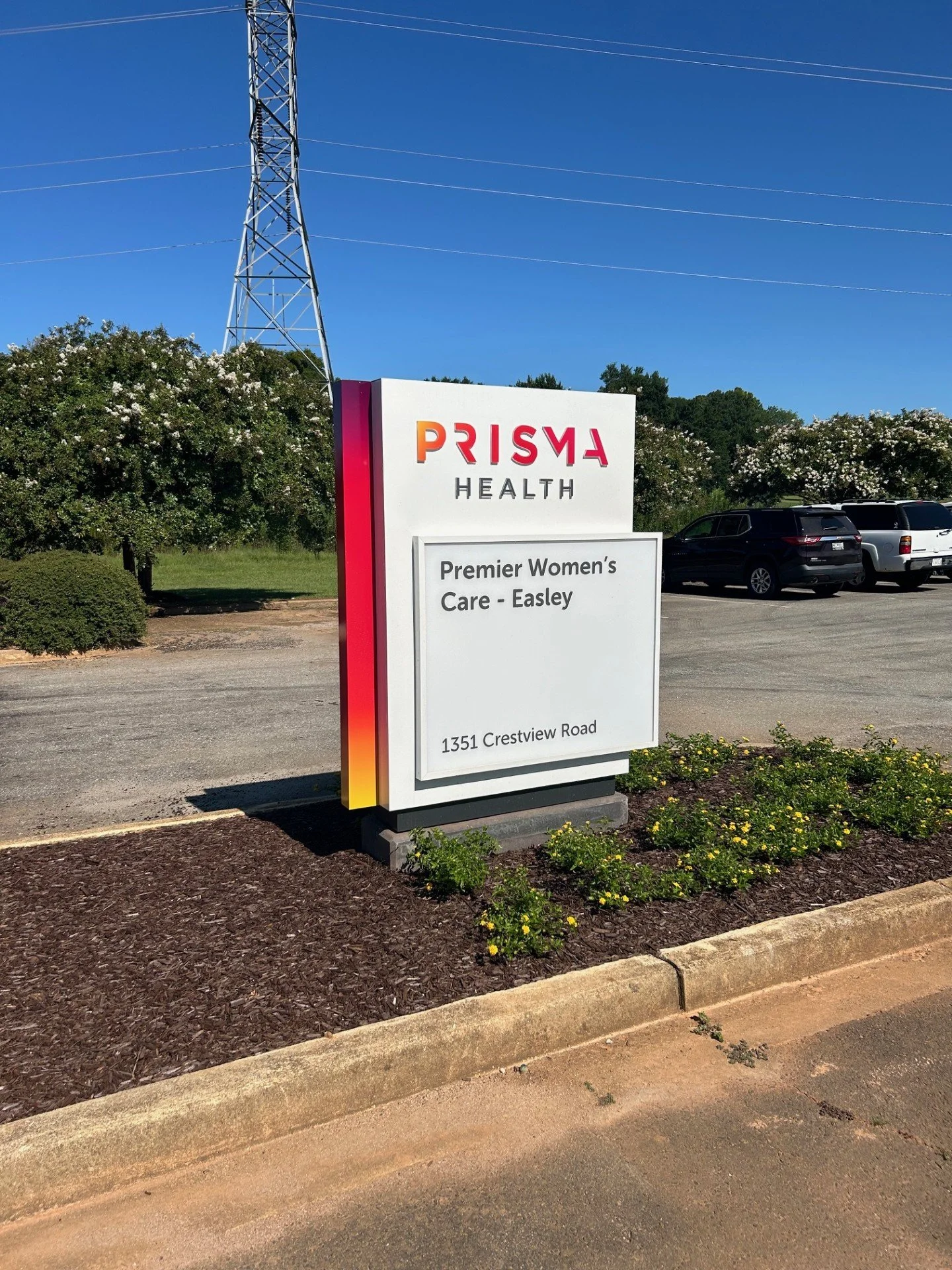 Sign for Prisma Health at 1351 Crestview Road, advertising premier women's care in Easley, with parked cars and greenery in the background.