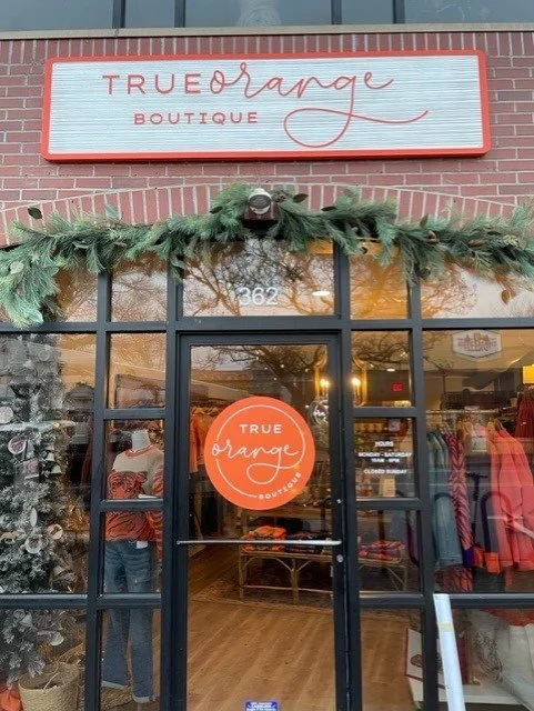 Storefront of True Orange Boutique decorated with Christmas garland, showing a glass door and windows with clothing displays inside.