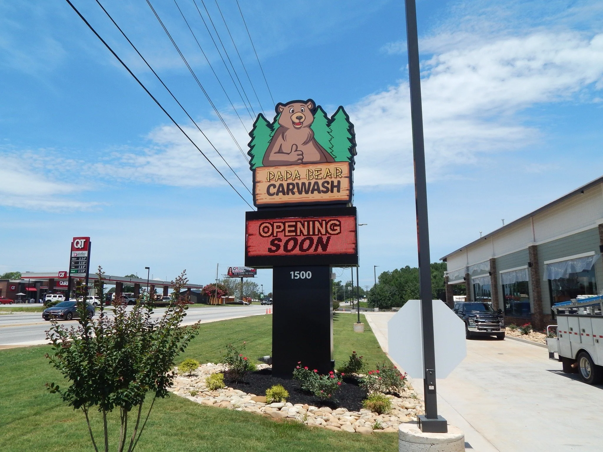 Sign for Papa Bear Carwash with bear cartoon giving a thumbs up, trees background, blue sky with some clouds, nearby gas station, cars parked in front of a building, and a sidewalk with landscaped area.