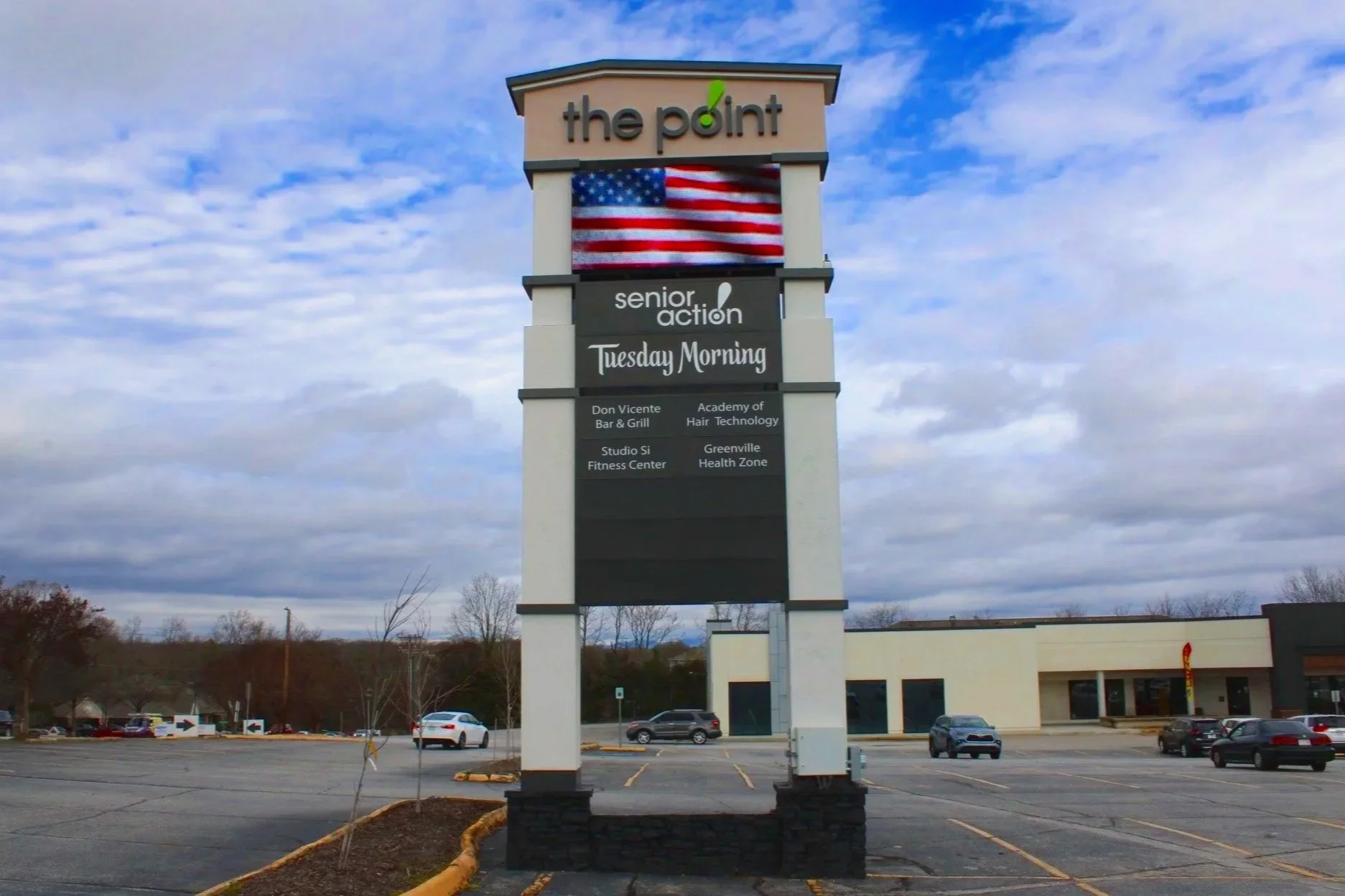 A parking lot with a large shopping center sign displaying 'the point,' a digital screen with an American flag, and businesses including senior action, Tuesday Morning, Don Vicente Bar & Grill, Academy of Hair Technology, Studio Si Fitness Center, and Greenville Health Zone, under a partly cloudy sky.