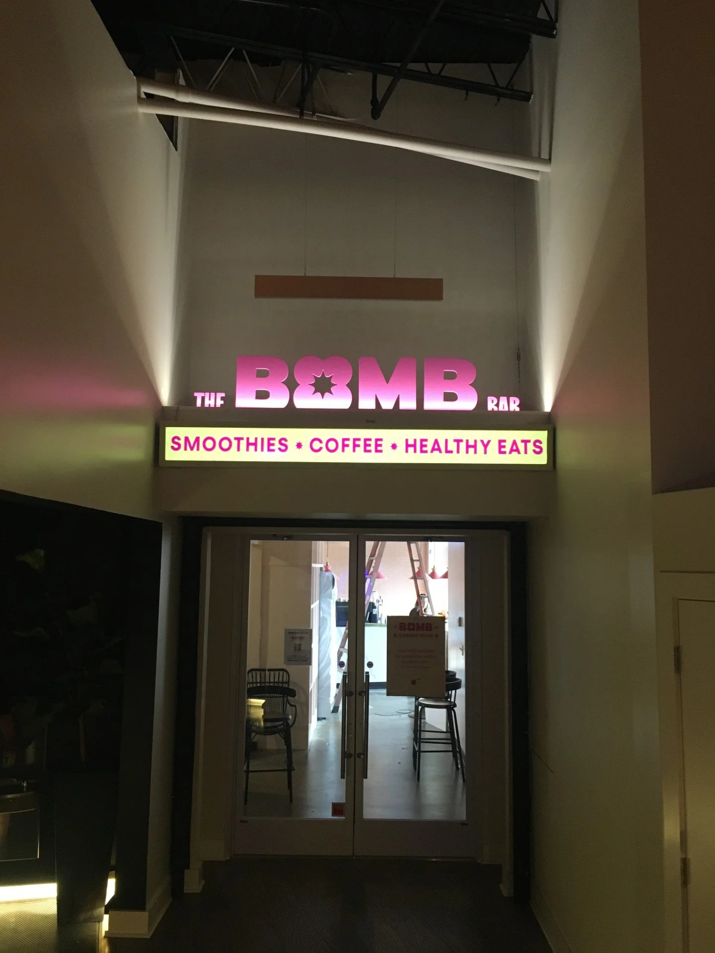 Entrance to a bar called 'The Bomb' with a pink illuminated sign. Below it, a white sign advertises smoothies, coffee, and healthy eats. The interior has chairs and restaurant equipment visible through glass doors.